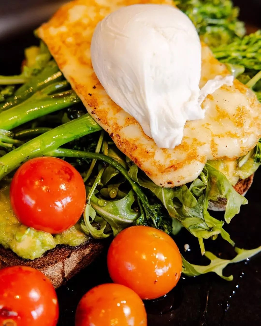 A close-up of our iconic Laika Stack - smashed avo, rocket, broccolini, halloumi, cherry tomato, a honey drizzle, a perfectly poached egg, all on toasted rye.