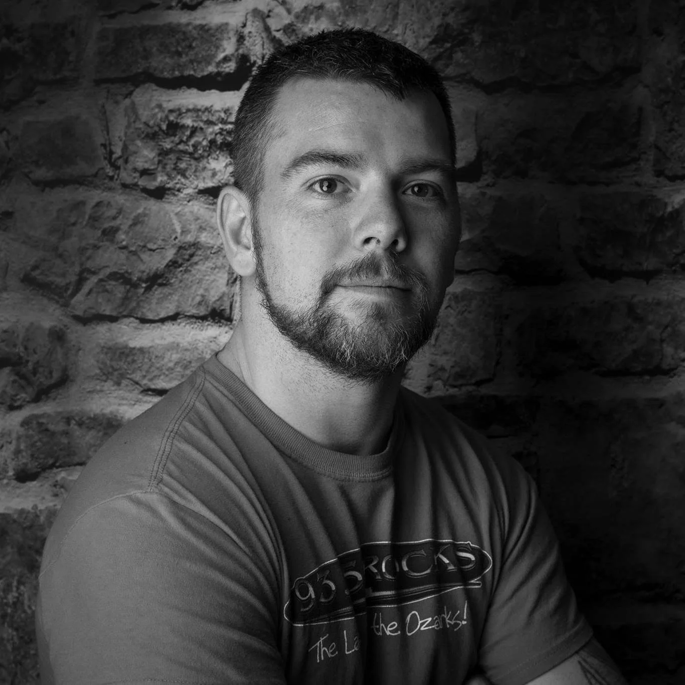 A man with a beard and short hair, wearing a T-shirt, sitting against a stone wall.