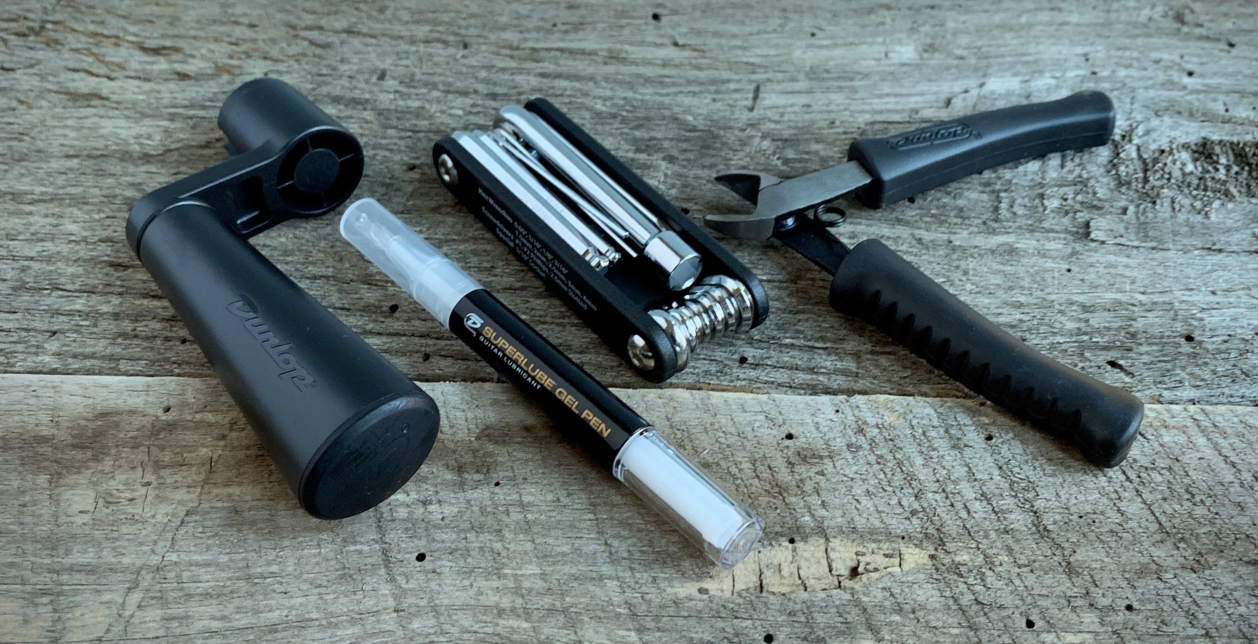 Guitar maintenance tools including a string winder, multitool, string cutter, and lubricant pen on a wooden surface.