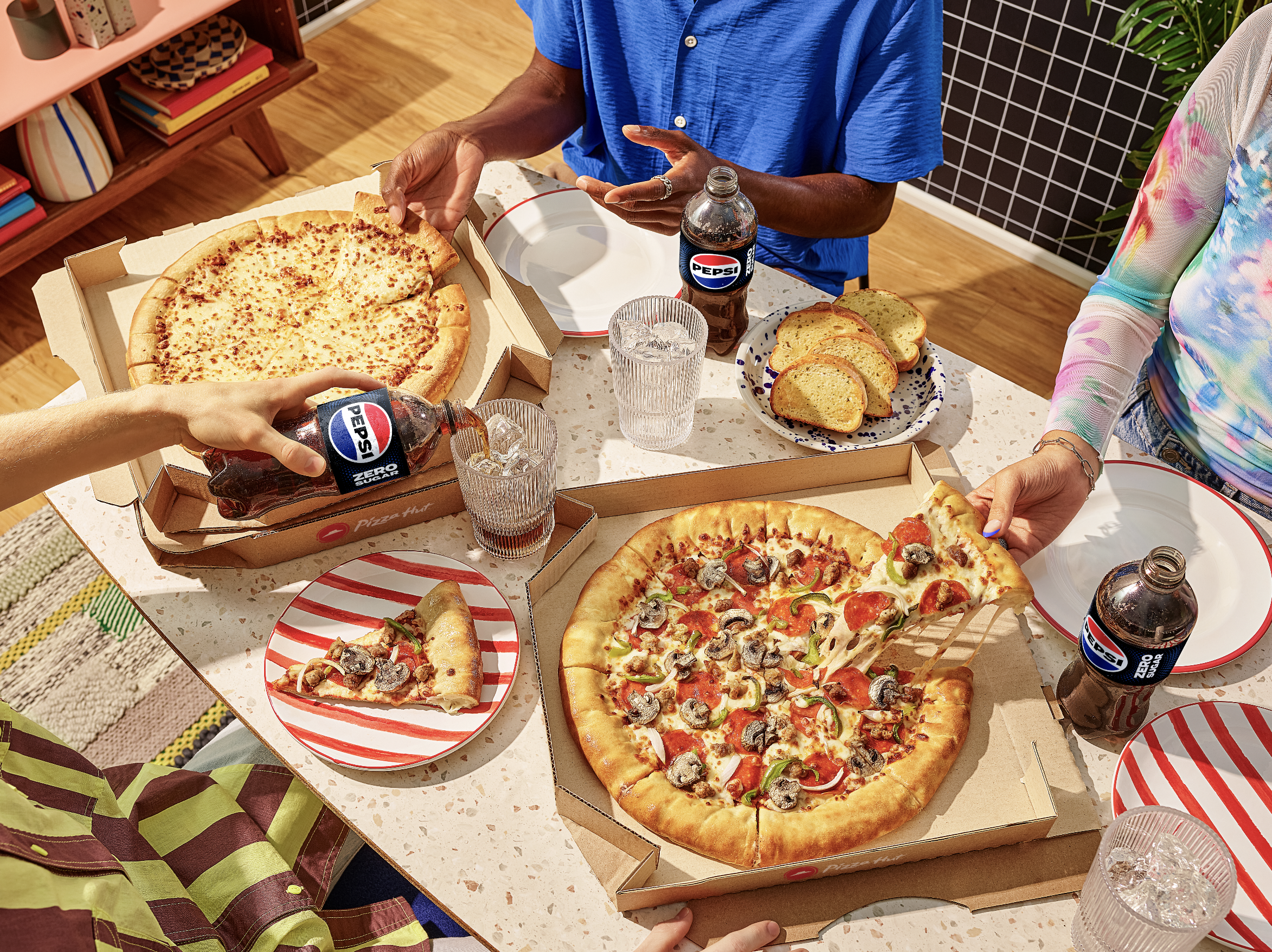 People sharing pizza and drinks at a table. There are two pizzas, slices of garlic bread, glasses of water, and bottles of Pepsi.