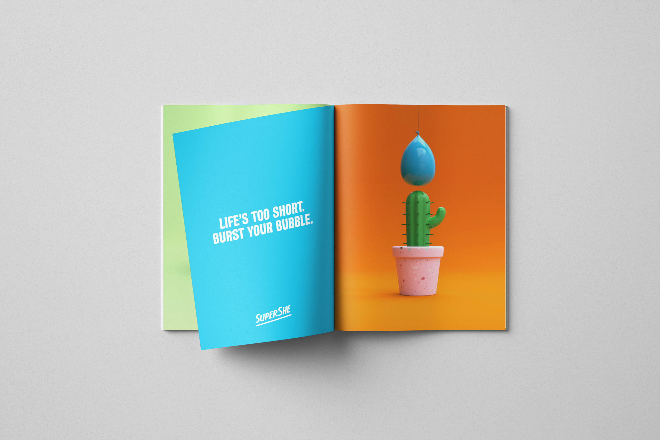 Open magazine with colorful pages: left page is blue with white text reading 'Life's too short, burst your bubble' and logo 'Supershe'; right page displays a cactus in a pink pot with a blue water balloon balanced on a string above it, set against an orange background.
