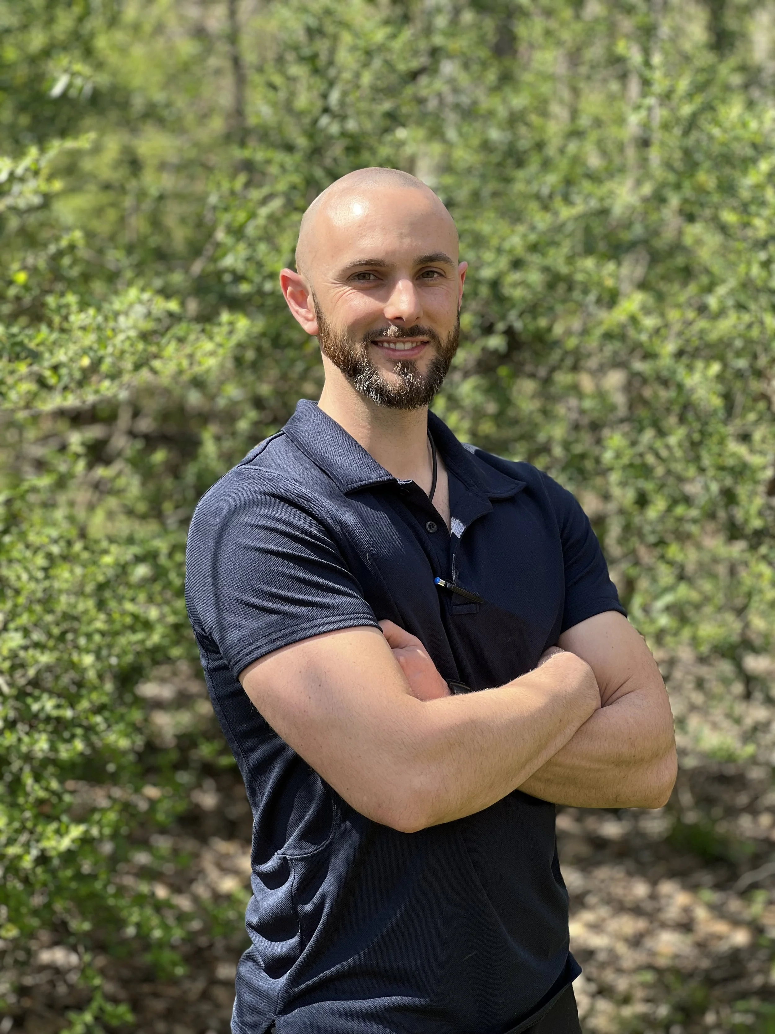 A man with a bald head and beard stands outdoors with his arms crossed, smiling, in a green wooded area.