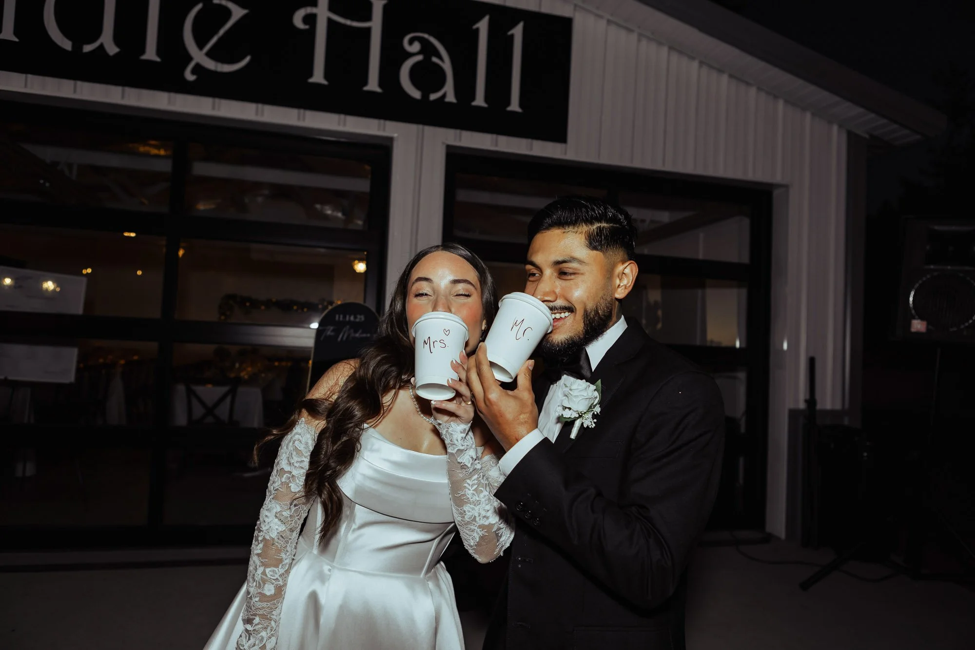 A newlywed couple, a woman in a white wedding dress with lace sleeves and a man in a black tuxedo, holding white cups labeled 'Mrs.' and 'Mr.' respectively, at a wedding reception.