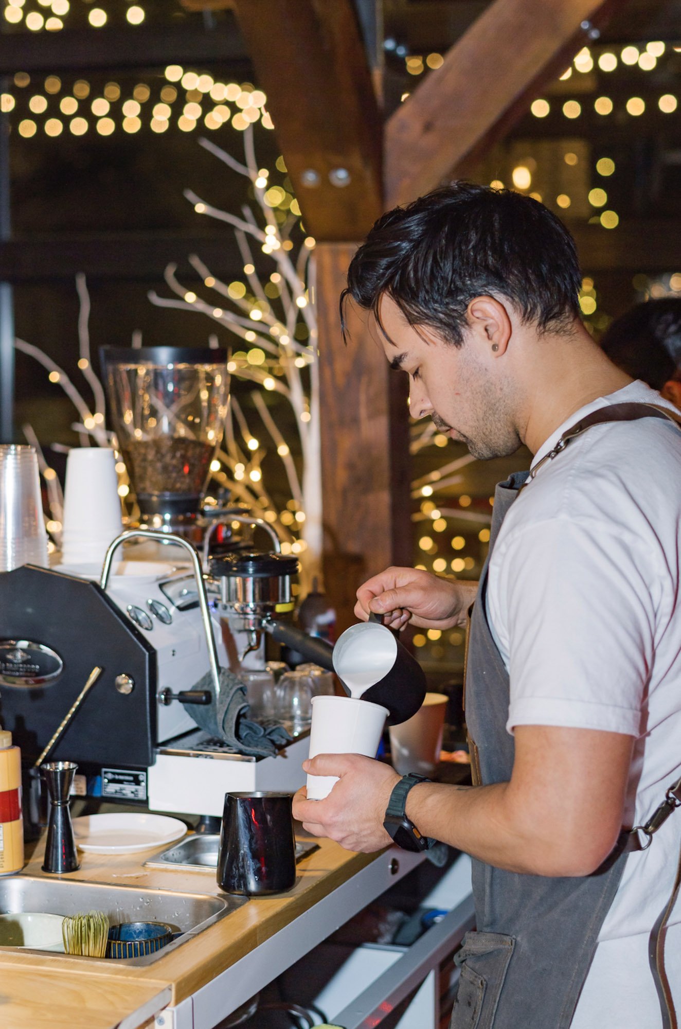 A man wearing a gray apron and wristwatch prepares coffee behind a counter with espresso machine and cups, decorated with string lights in the background.