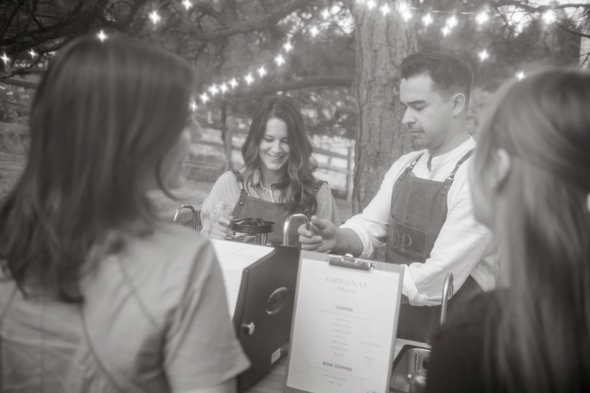 A group of four people gathered around a coffee stand outdoors, smiling and engaged in conversation. The scene appears to be in a park with trees and string lights overhead.