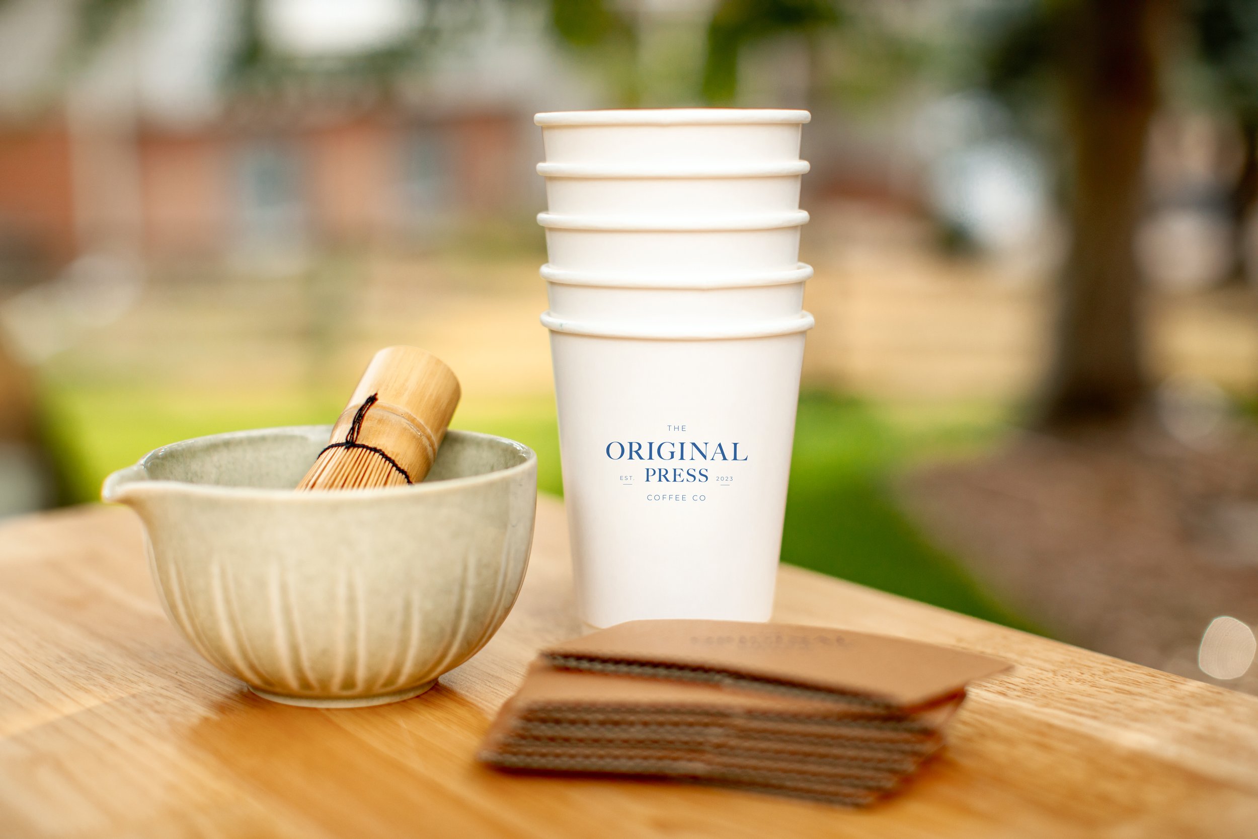 Stacked white coffee cups with a logo reading 'The Original Press Coffee Co' on a wooden table outdoors, with a small bowl containing a bamboo whisk nearby, blurring of background foliage.