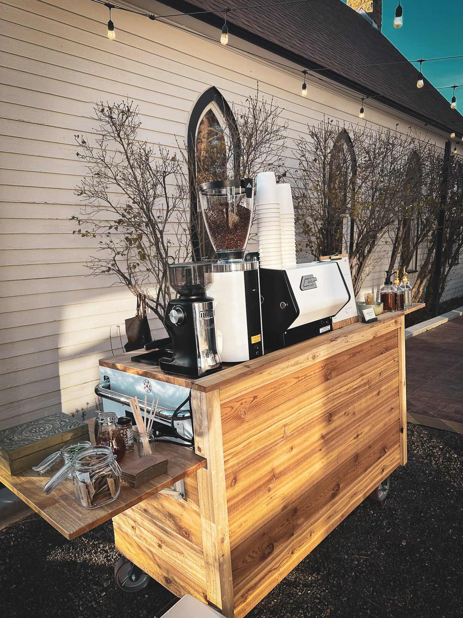 A coffee station outdoors on a wooden cart with a coffee grinder, cups, syrups, and jars of flavorings, with leafless bushes and string lights overhead.