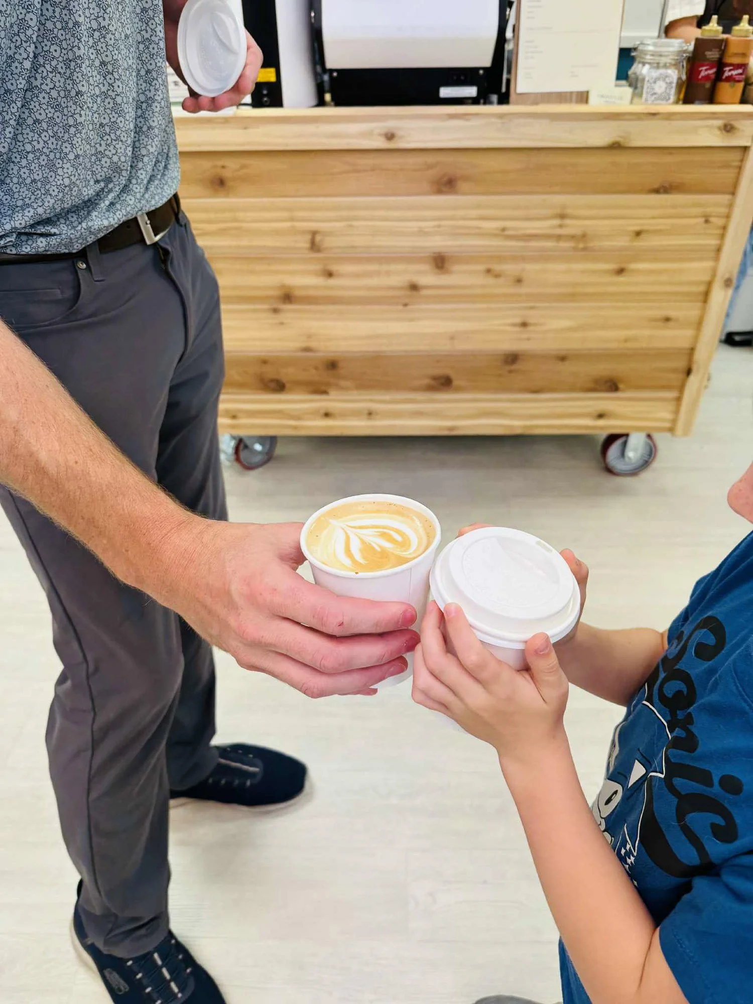 Two people exchanging cups of coffee inside a cafe or coffee shop. One person is wearing a gray polo shirt with a dotted pattern, gray pants, and sneakers. The other person is wearing a blue T-shirt with a design on the sleeve. The background shows a wooden counter with various bottled drinks and a cash register.