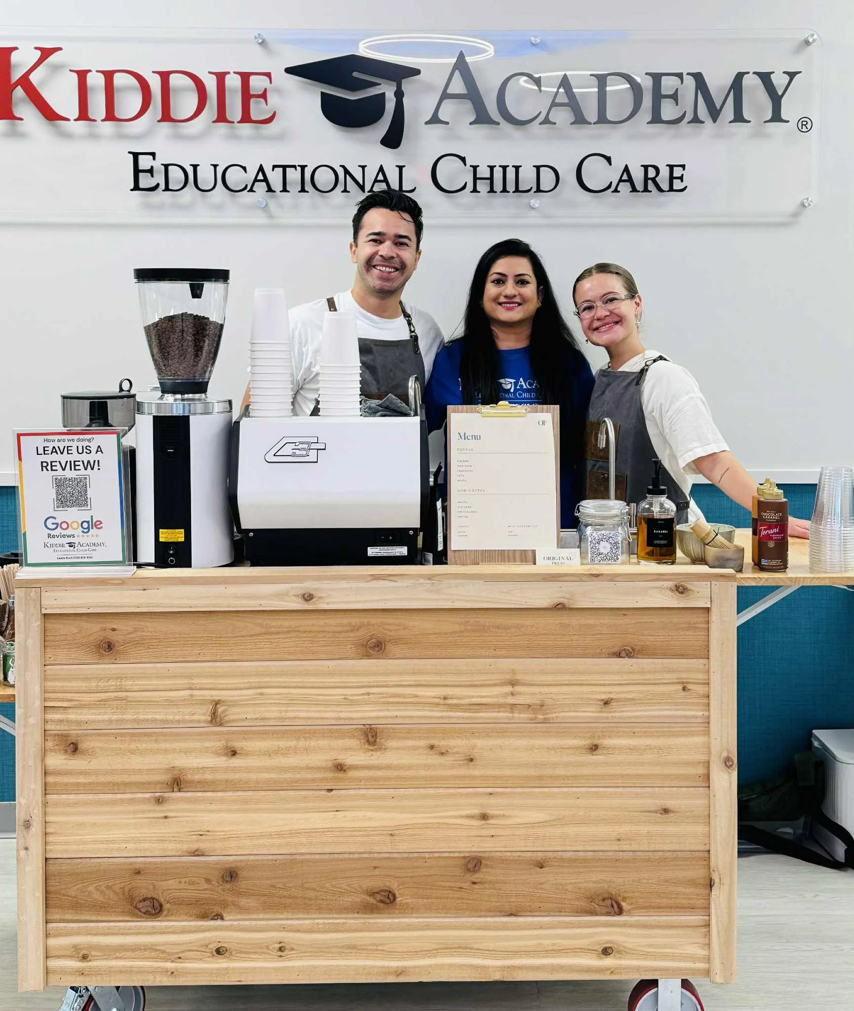 Image of a wooden counter with coffee-making equipment, a menu, and jars, in front of a sign that reads 'KIDDIE ACADEMY EDUCATIONAL CHILD CARE.' Three staff members, two women and one man, are smiling behind the counter.
