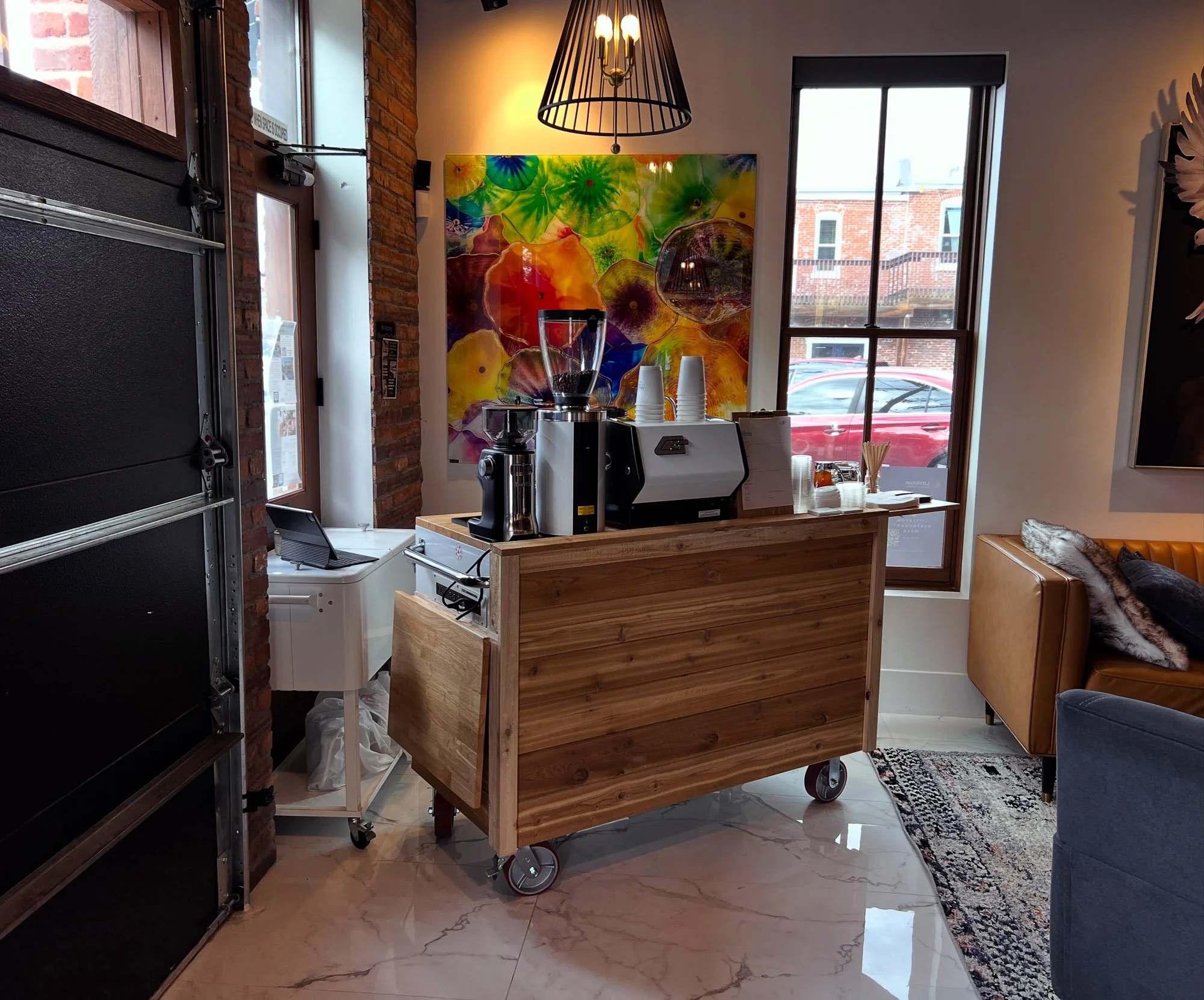 Interior of a cafe or coffee shop with a wood cart, coffee machines, cups, and a colorful abstract wall art behind. Large windows showing parked cars and brick buildings outside.