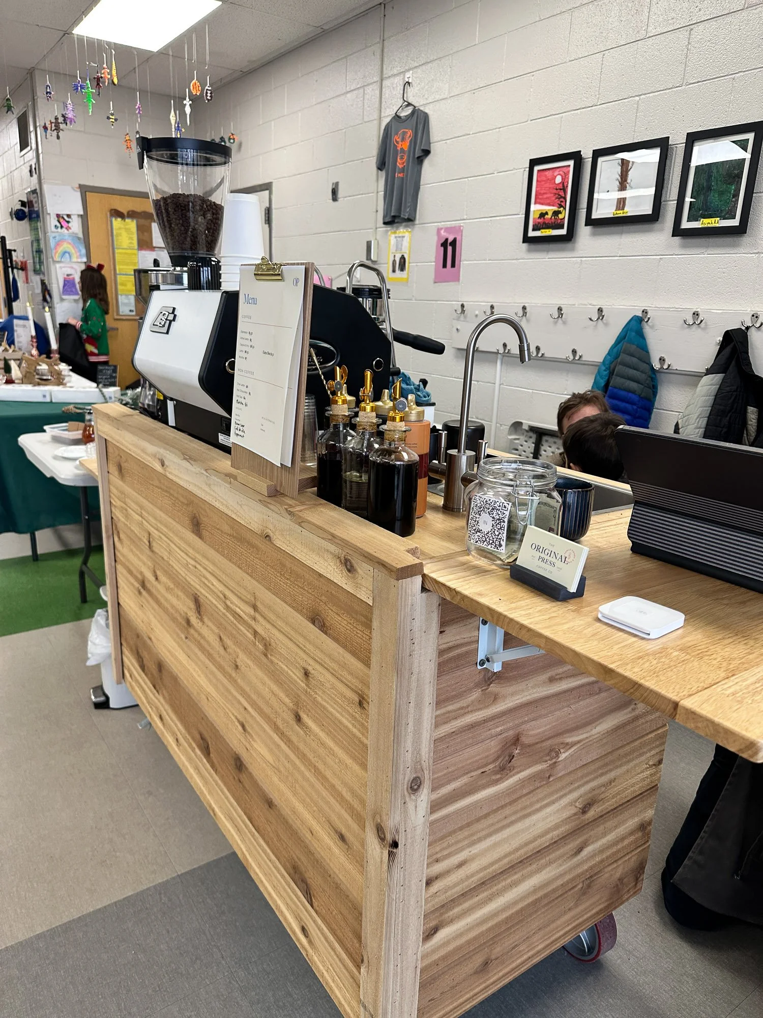A wooden counter with various items on top, including syrup bottles, a coffee machine, a menu, and a jar with a QR code. Behind the counter, a white brick wall has framed pictures, hooks with coats, and a gray T-shirt hanging.