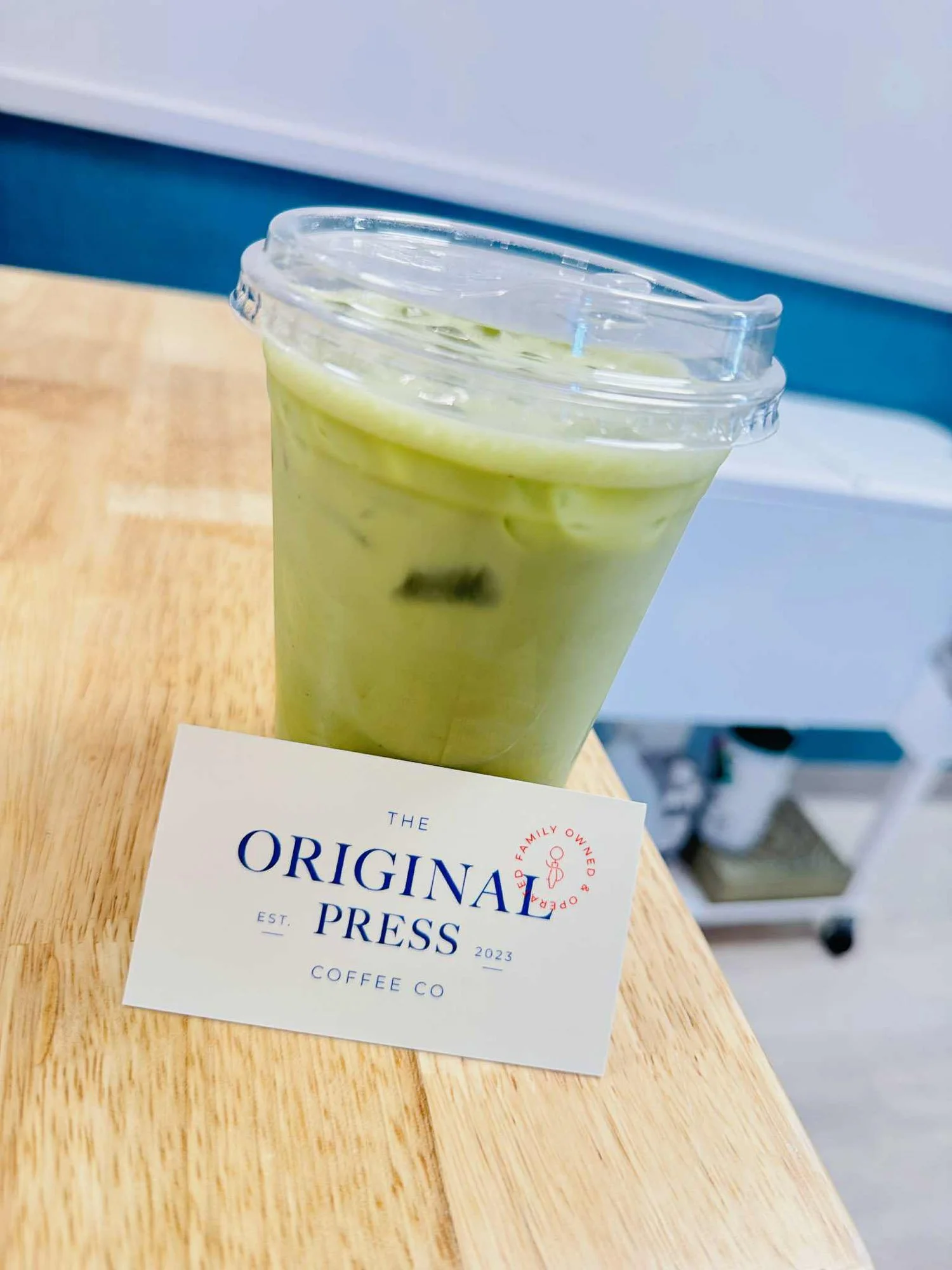 A clear plastic cup filled with a green-colored iced beverage, placed on a wooden surface, with a white paper card in front that reads 'The Original Press' and 'Coffee Co,' with a red circular stamp indicating that it is family owned and operated.