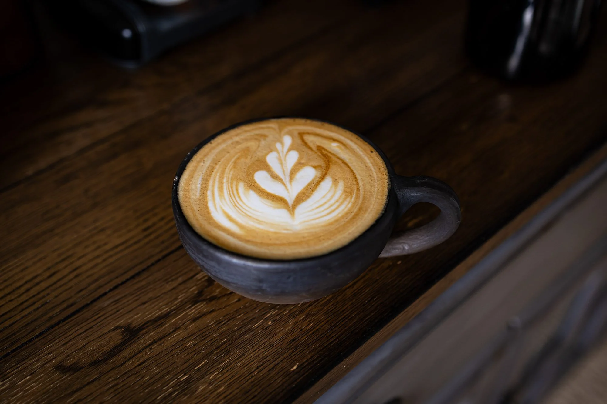 Black mug with latte art on a wooden table, with a black espresso machine and a black cup in the background.