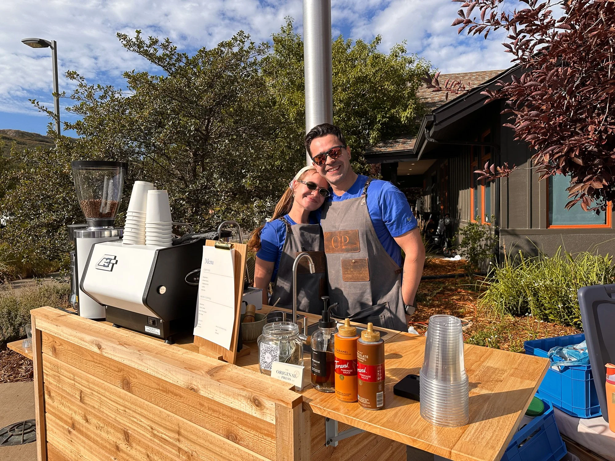 A man and woman standing behind a wooden coffee stand outdoors. The man is wearing sunglasses and an apron, and the woman is leaning on him. The stand has coffee equipment, syrups, and cups, with a menu and a QR code. Behind them are trees, a building, and a partly cloudy sky.