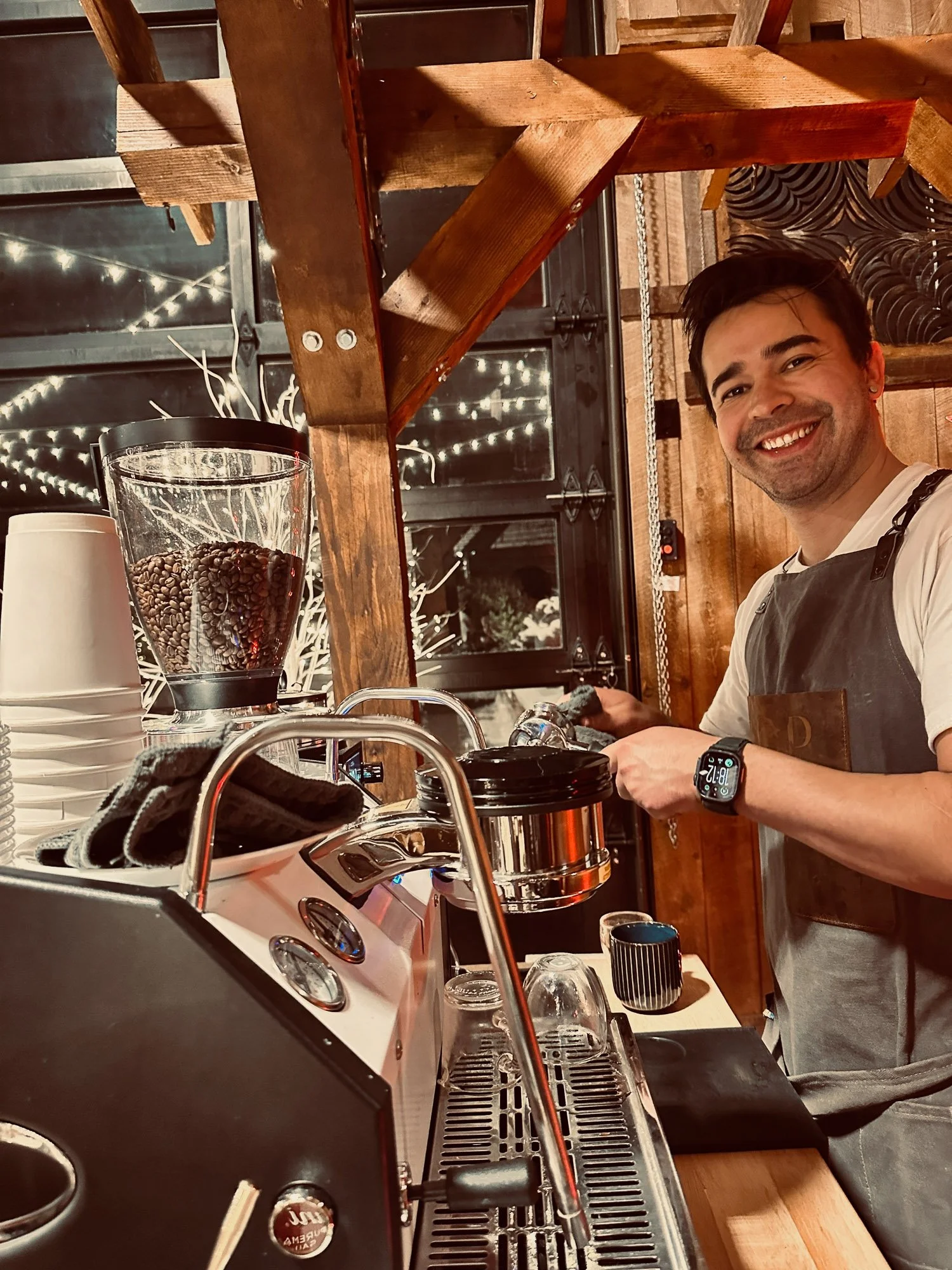 A smiling man in a white shirt and gray apron working behind a coffee bar with espresso machine, coffee grinder with beans, cups, and barista tools, in a wooden cafe setting.