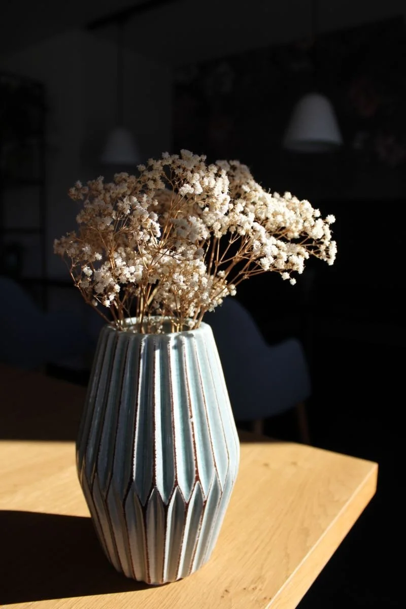 Blumen in einer gestreiften Vase auf einem Holztisch, im Hintergrund dunkler Raum mit Deckenlampen.