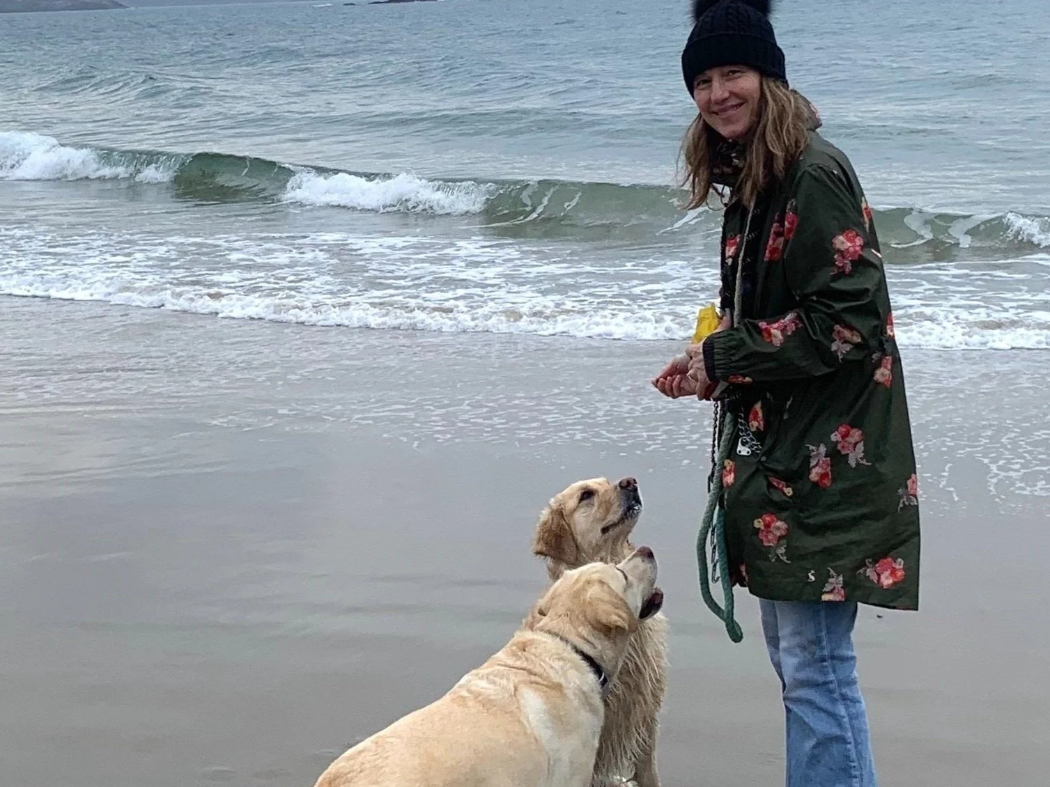 Person wearing a green floral coat and black beanie walking two dogs on a beach with waves in the background.