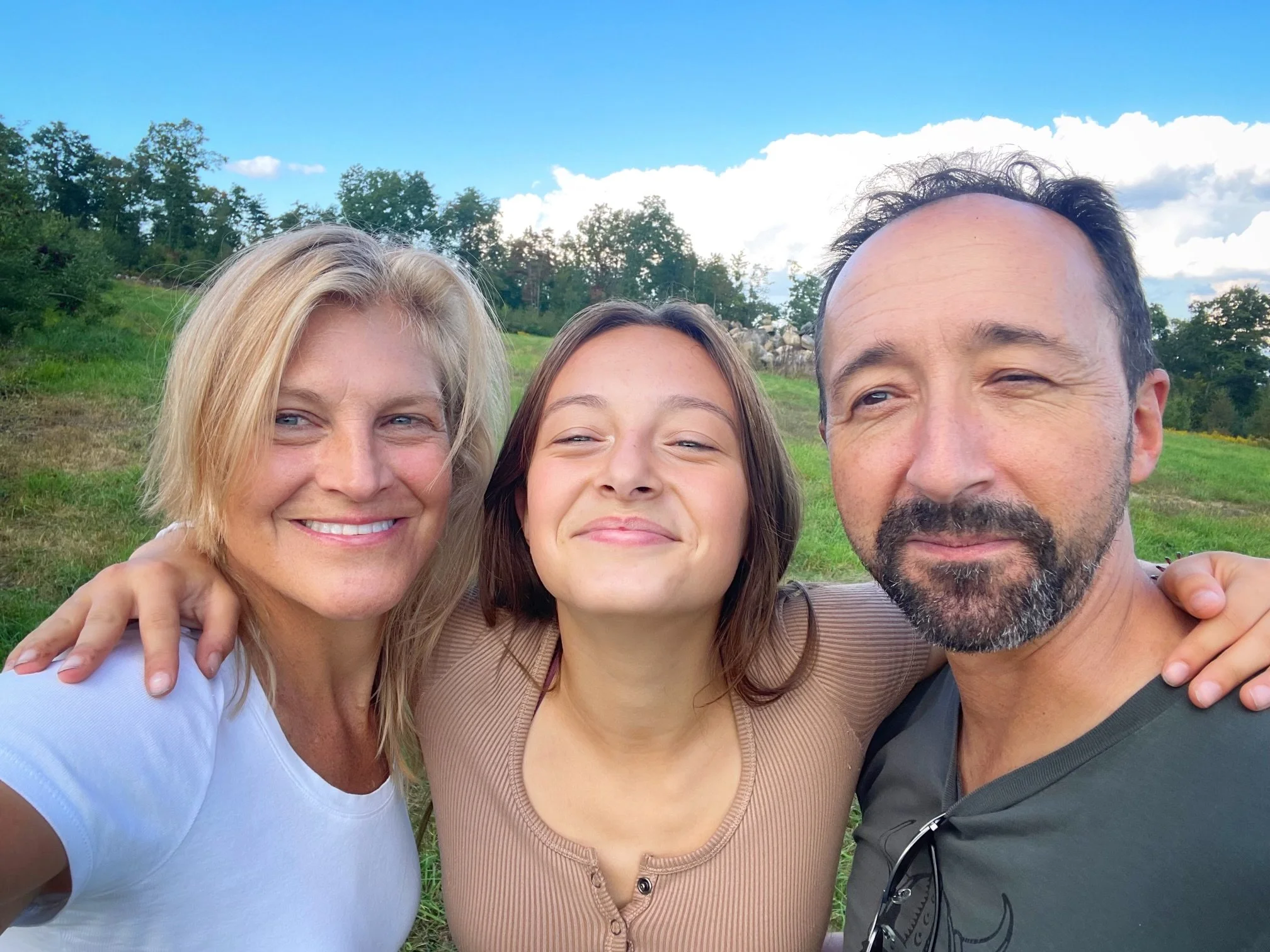 Three people smiling in a grassy field with trees and a blue sky in the background.