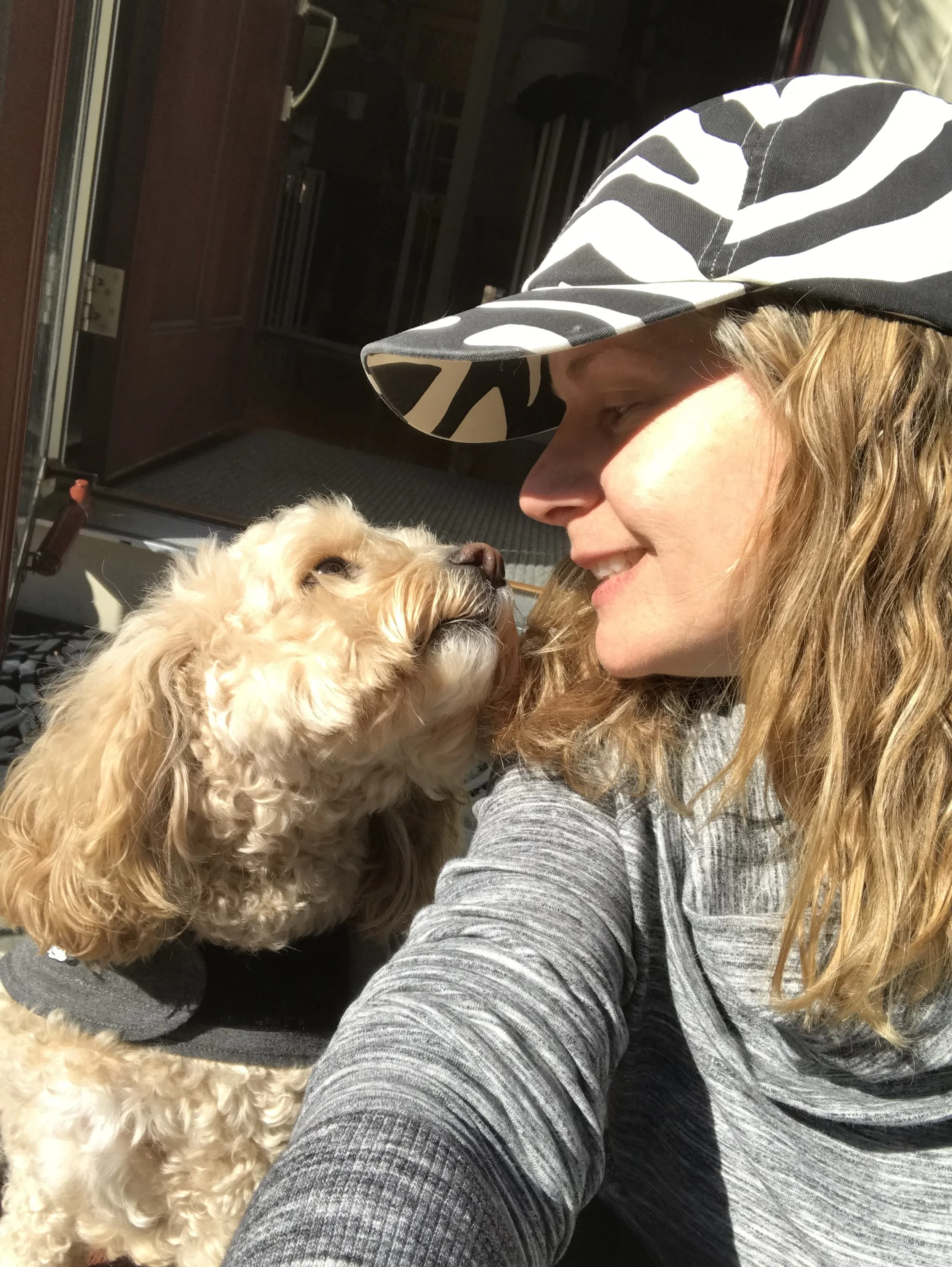 Woman wearing a zebra-print visor smiling at a cream-colored dog.