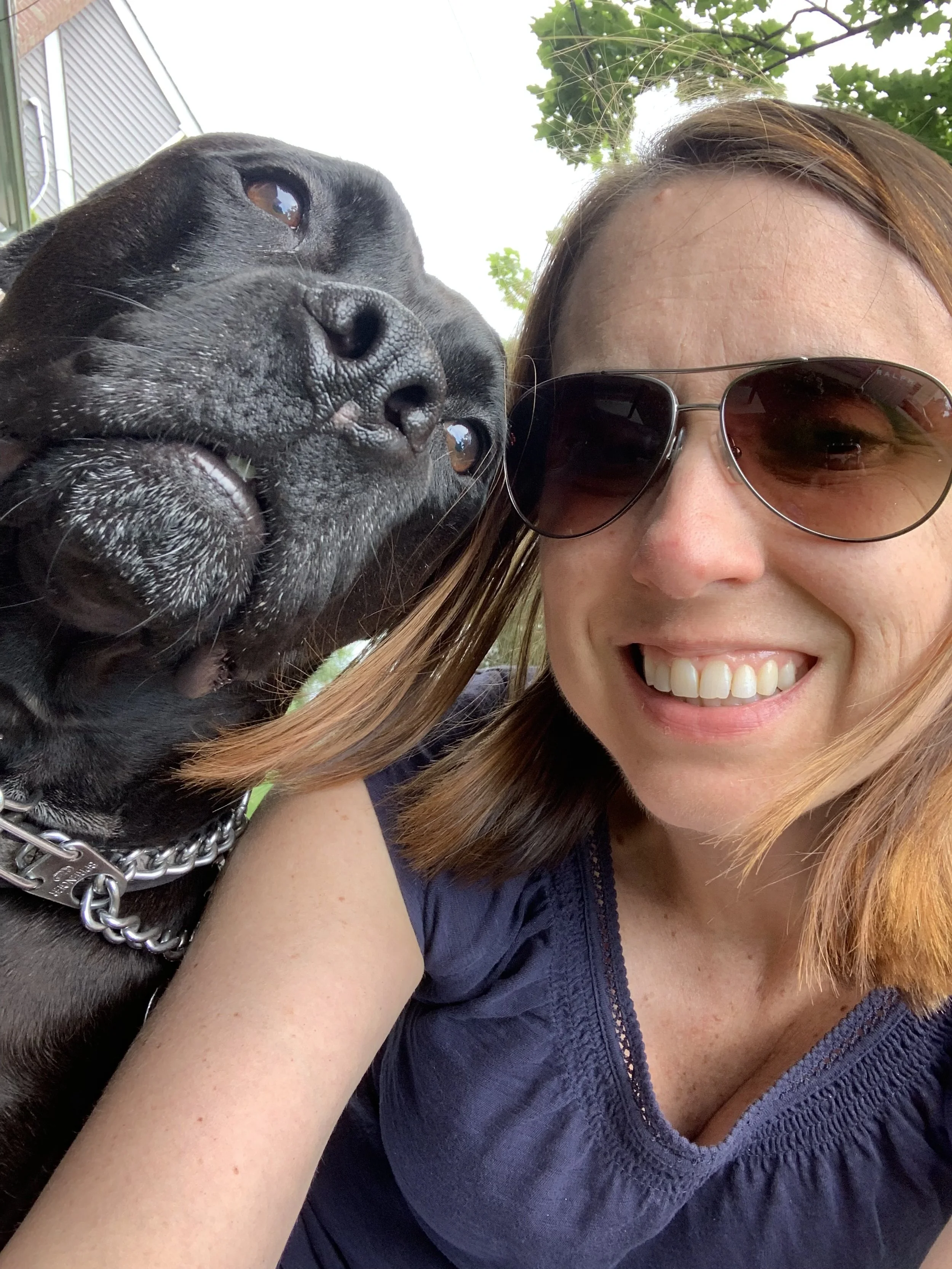 Woman wearing sunglasses smiling with a black dog, both in close-up, outdoors.
