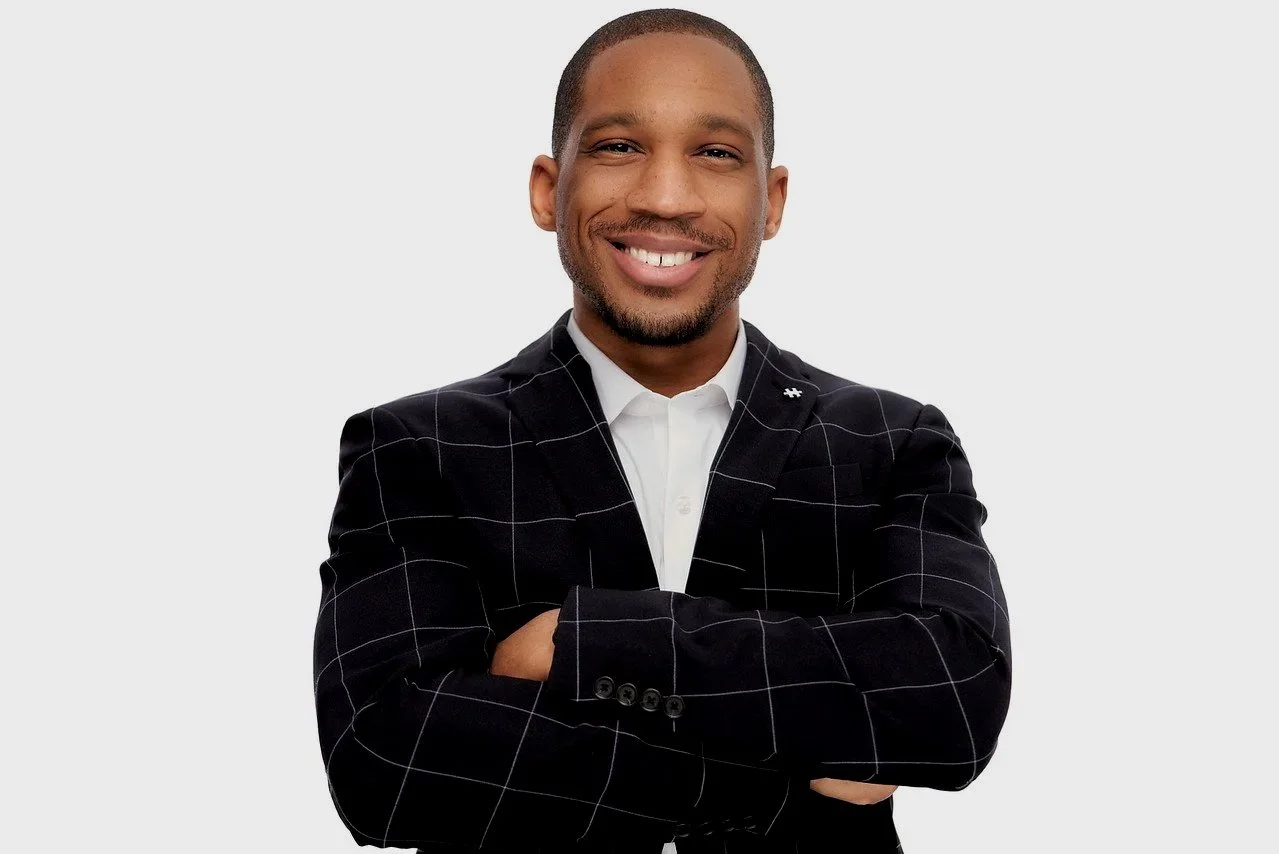 A smiling man with crossed arms wearing a black checked blazer and white shirt against a light gray background.