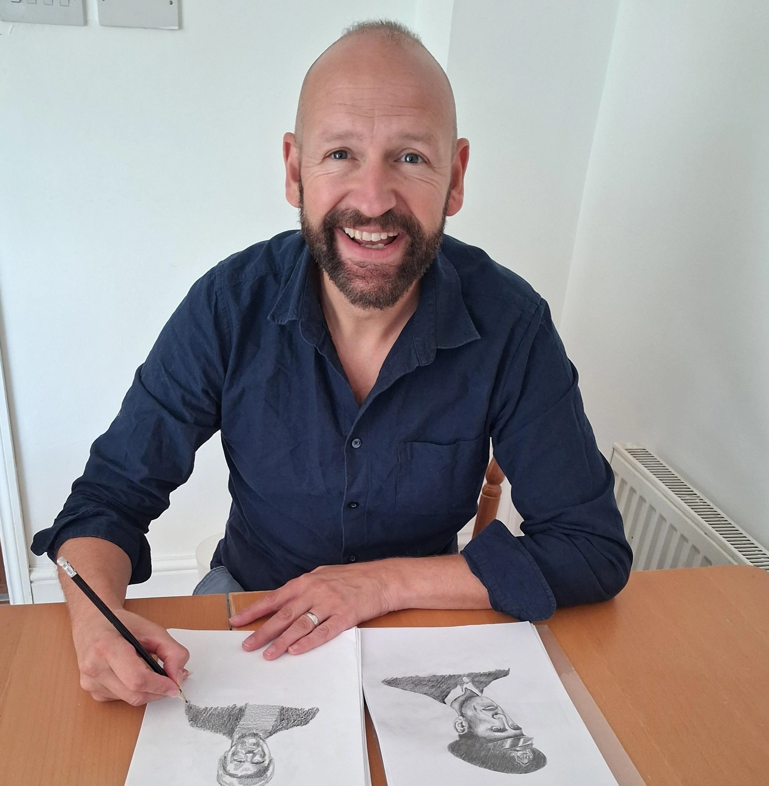 Christopher James with a beard and bald head smiling, sitting at a wooden table, drawing pencil sketches of members of the crew with a pencil on paper.
