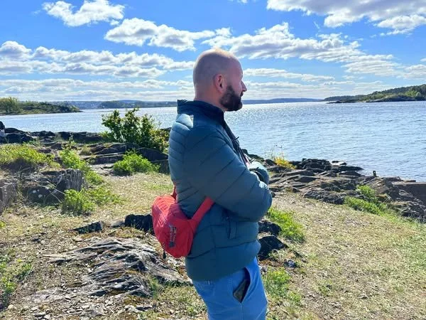 A man with a beard and shaved head wearing a blue jacket and red backpack stands on a rocky shoreline with arms crossed, facing a body of water under a partly cloudy sky.