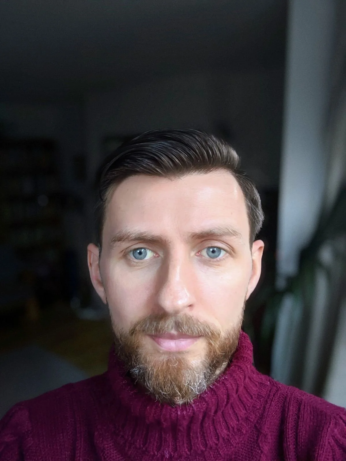 Close-up selfie of tom McLaughlin with blue eyes, short dark hair, and a beard, wearing a maroon cable-knit turtleneck sweater, indoors with a dark background.