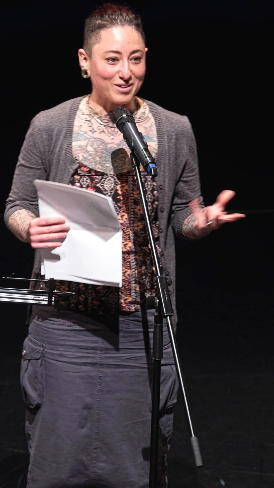 A woman with short hair and tattoos, wearing a patterned shirt and a gray cardigan, standing at a microphone on a black background, holding papers and gesturing with her left hand.