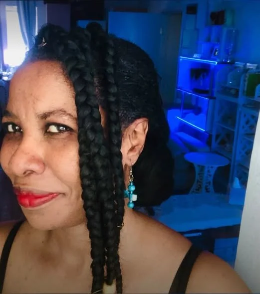 A woman with dark braided hair and earrings, smiling in a room with blue lighting.
