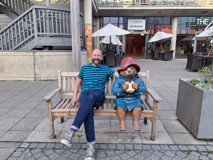A man sitting on a wooden bench next to a bear statue dressed in a blue coat and red hat, holding a slice of bread. The setting appears to be an outdoor shopping or dining area with umbrellas and storefronts in the background.