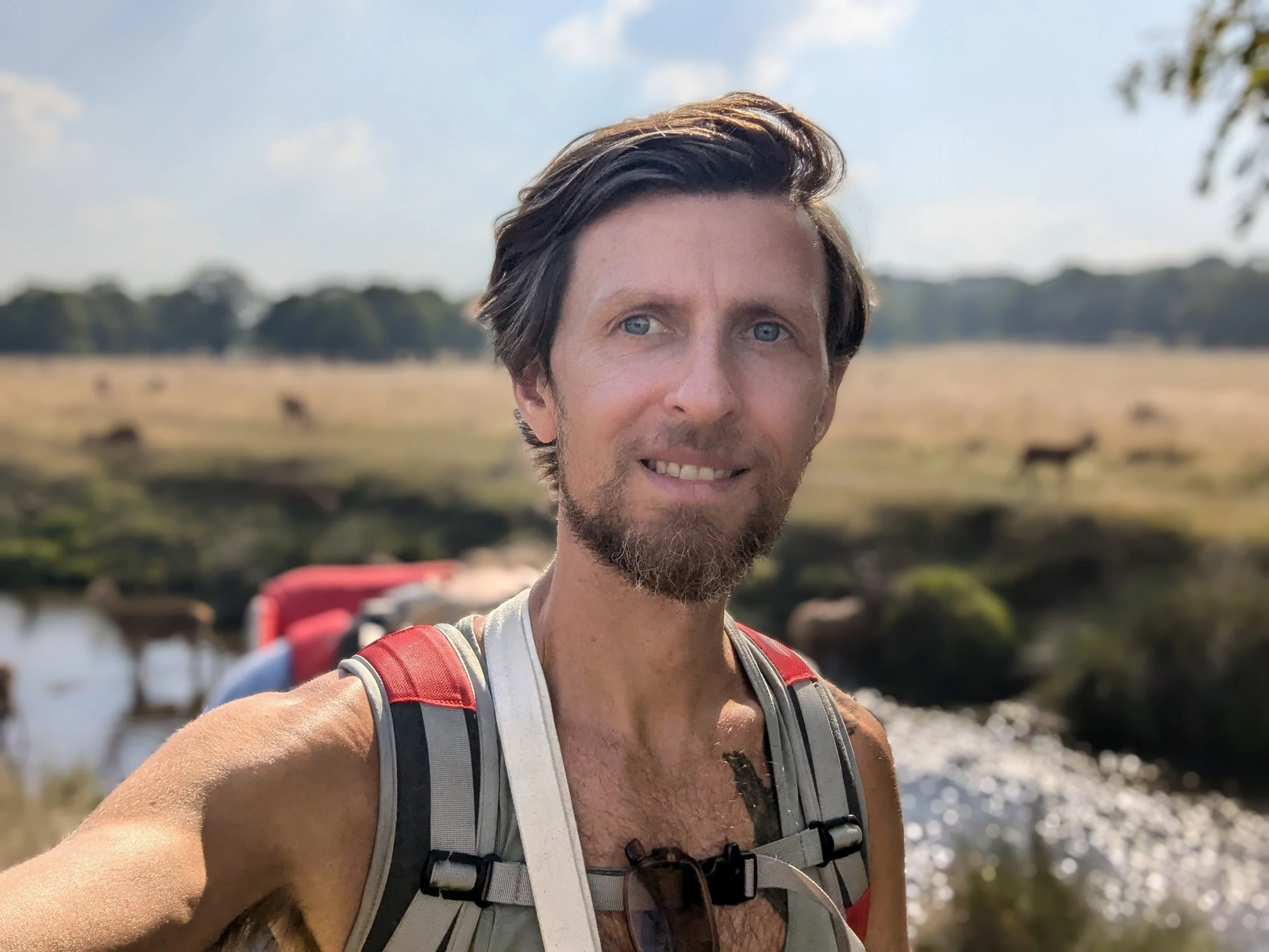 Tom McLaughlin, with brown hair and a beard, taking a selfie outdoors in a natural setting with a body of water, trees, and a blue sky with clouds in the background.