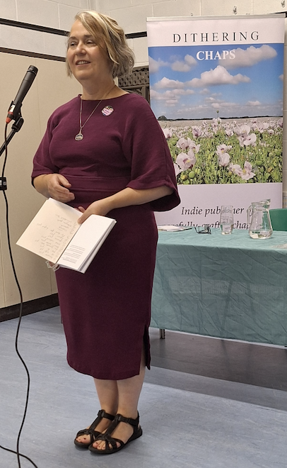 A woman in a purple dress speaking at a microphone, holding a book, with a banner and table behind her at an indoor event.