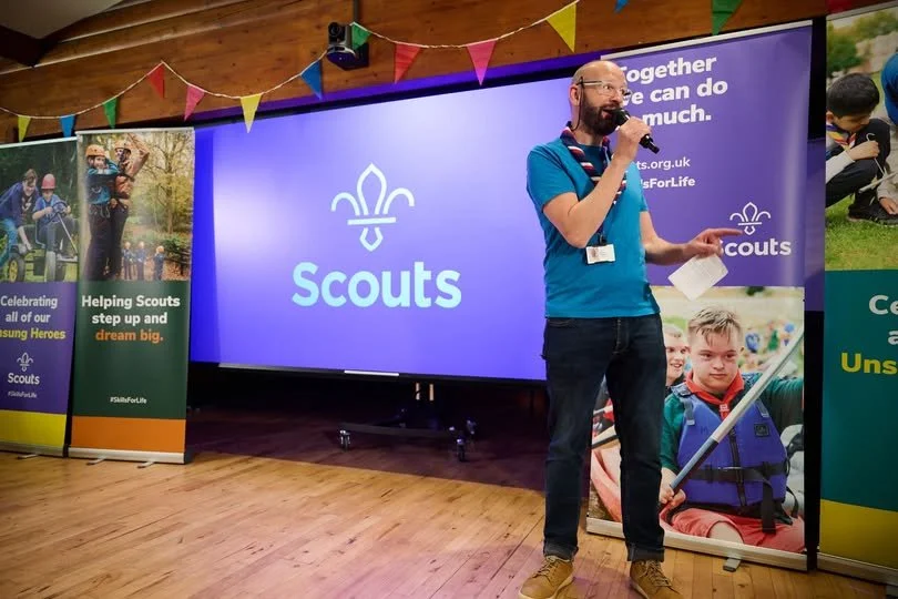 A man holding a microphone and a piece of paper standing to the right of a large screen displaying the Scouts logo. The background shows colorful bunting and promotional banners for Scouts.