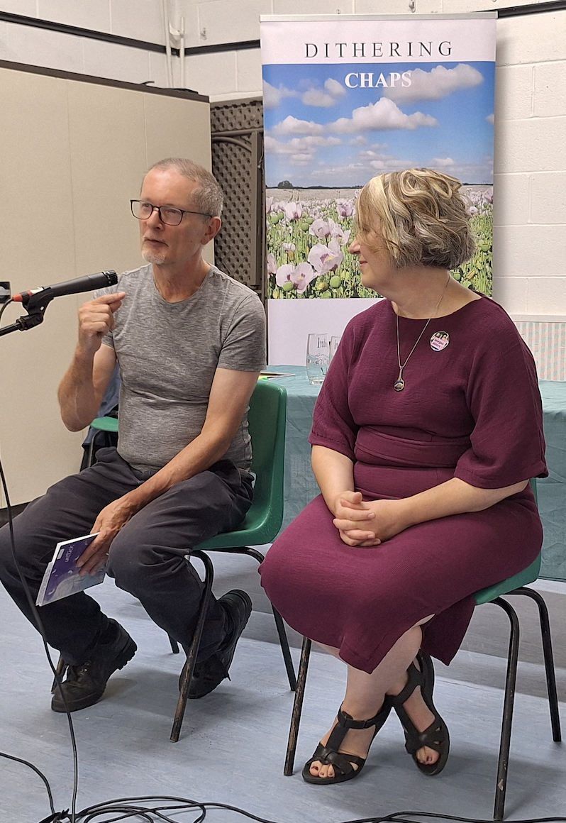 Two people sitting on chairs, engaged in a discussion with a microphone. The background includes a Dithering Chaps banner and a table with glasses.