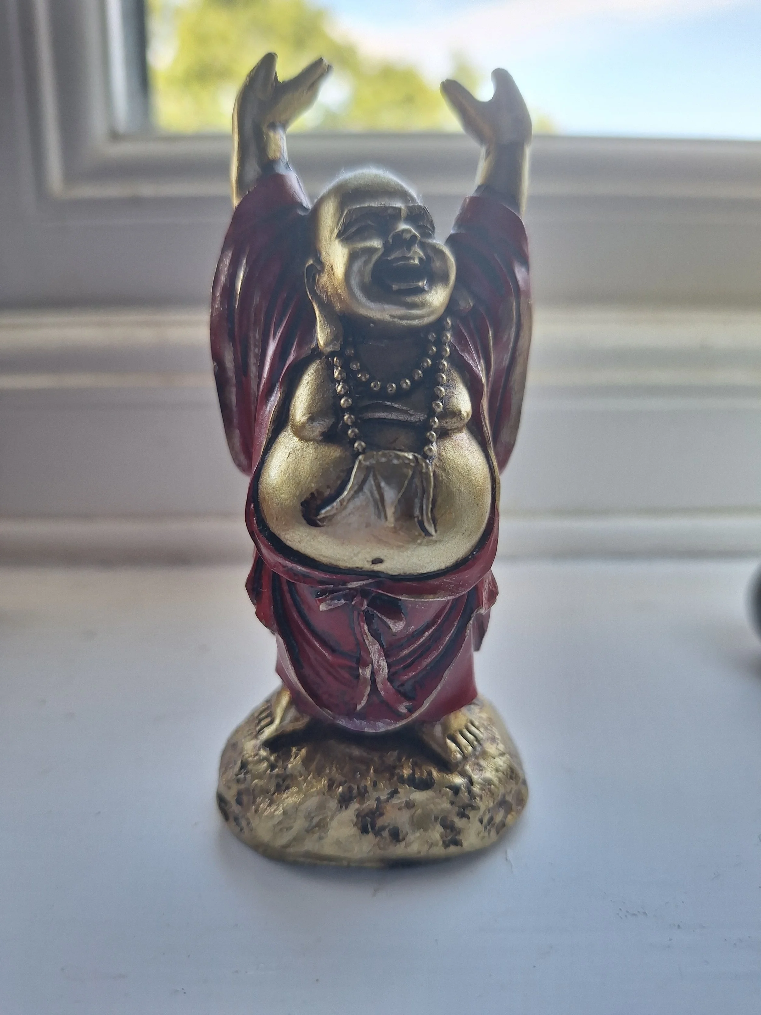 A figurine of a laughing, smiling Buddha with arms raised, dressed in a red robe with gold accents, standing on a textured base.