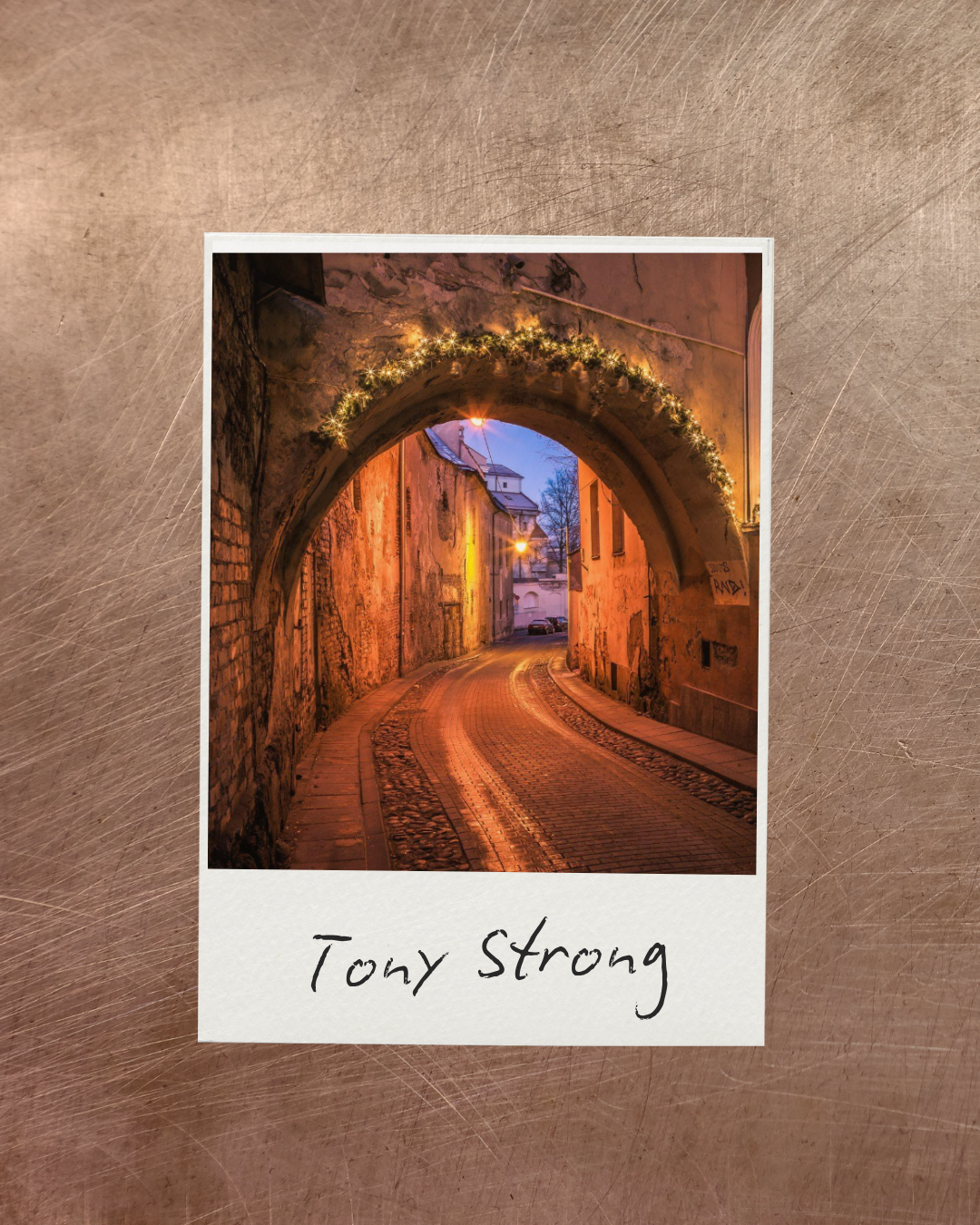 A nighttime photo of a narrow cobblestone street seen through an archway decorated with string lights, with buildings on either side and a faintly lit sky in the background and the name Tony Strong