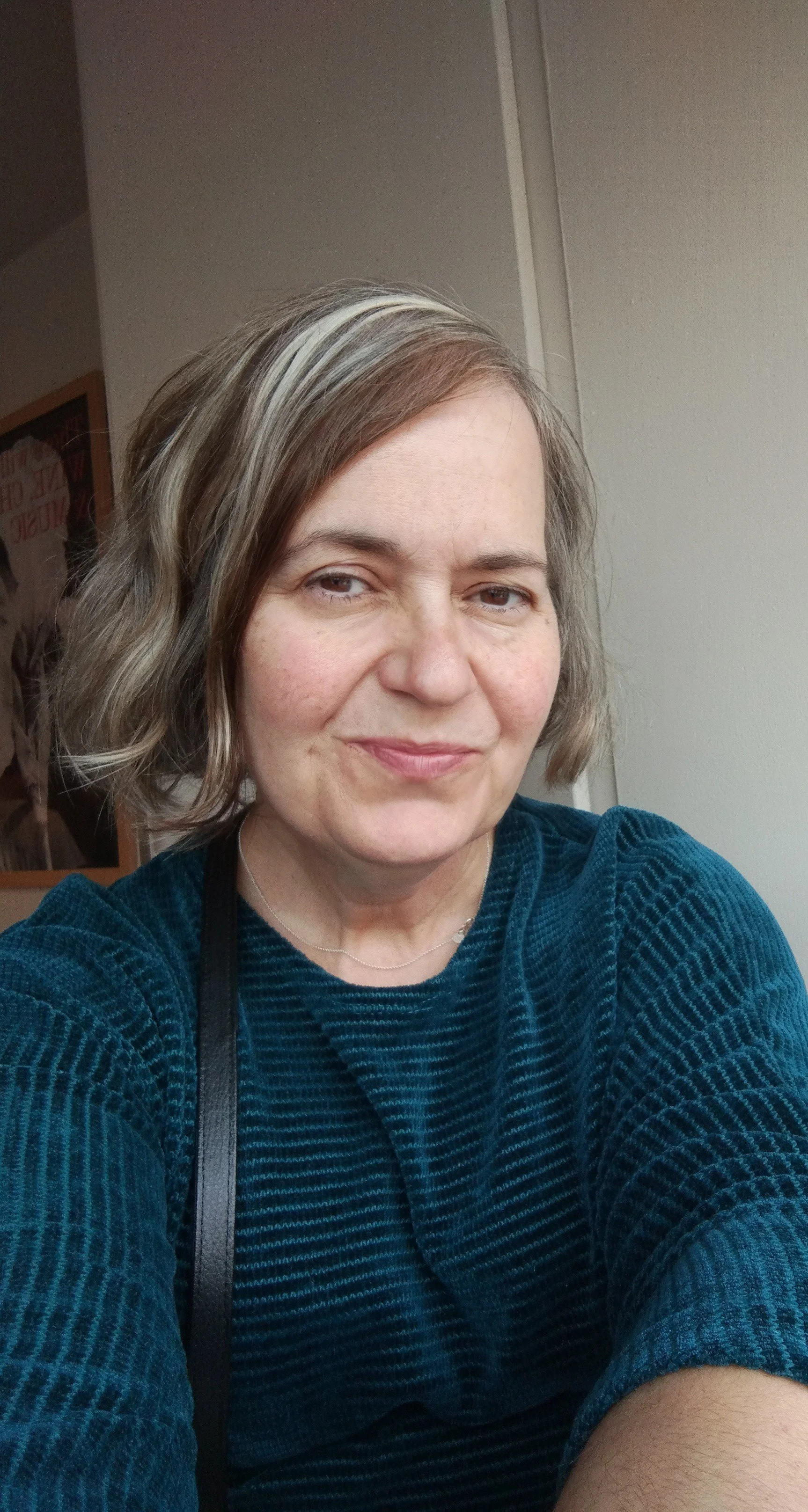 A woman taking a selfie, wearing a blue textured sweater, with short wavy hair and a slight smile in an indoor setting.