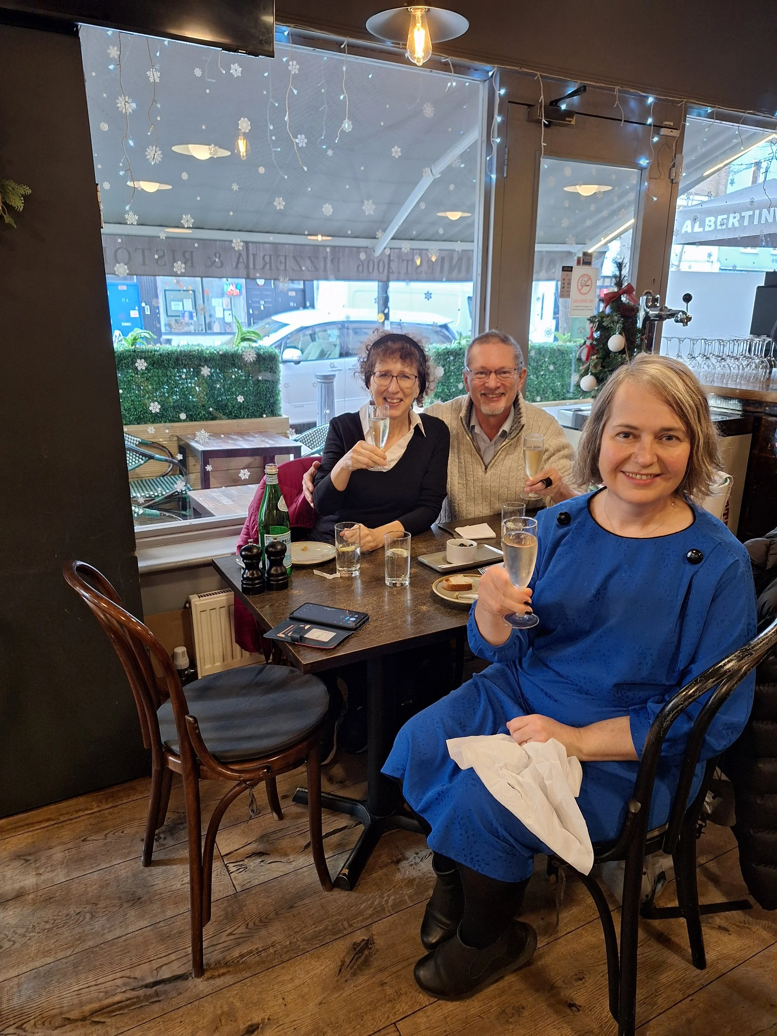 Three people sitting at a restaurant table celebrating, with champagne glasses raised. The setting has holiday decorations including snowflakes on the window and a small Christmas tree. The group is smiling and appears to be enjoying themselves.