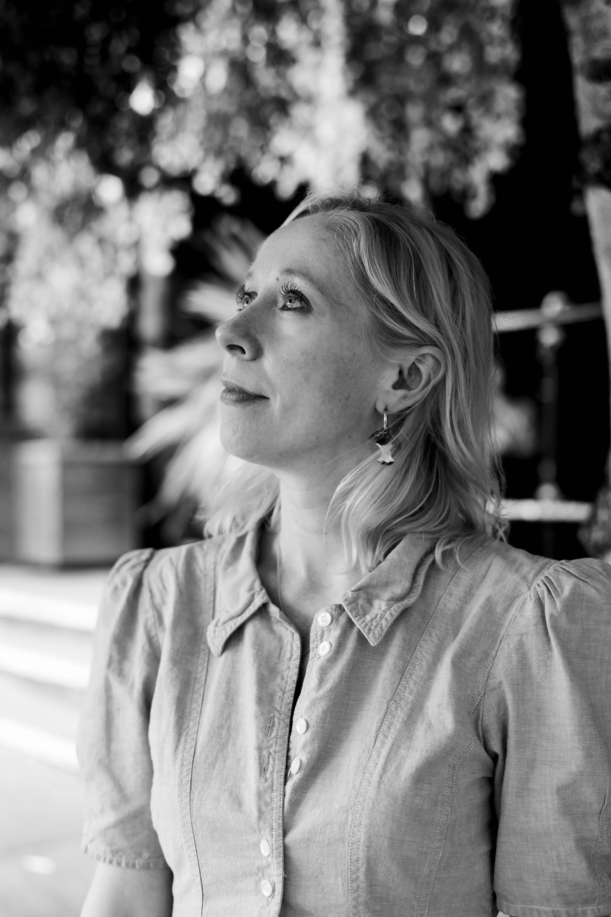 Black and white photo of Victoria Spires with blonde hair, wearing a button-up shirt, looking upward with sunlight on her face, and trees with blurred bokeh in the background.