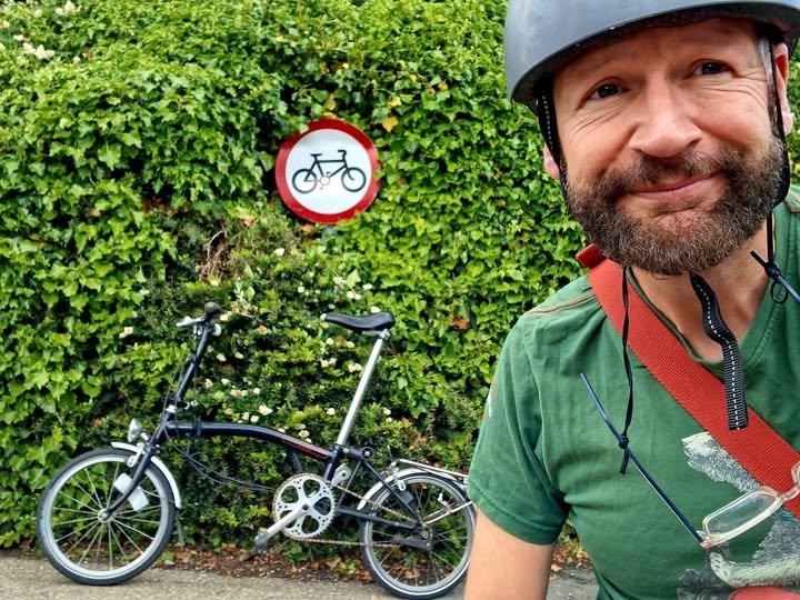 A man with a bicycle helmet smiling in front of a shrub wall and a sign indicating bicycles are not allowed, with a small folding bicycle parked on the ground beside him.