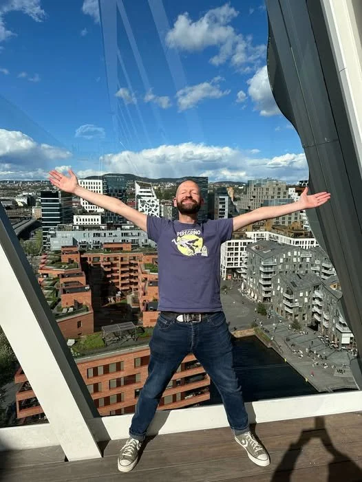 A smiling man standing on a balcony with arms outstretched, overlooking a cityscape with modern buildings and a blue sky with clouds.