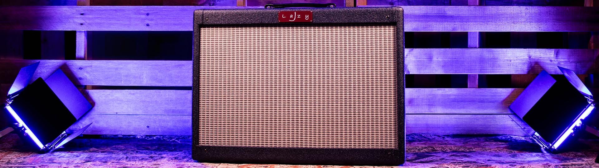 A guitar amplifier is positioned in front of a wooden pallet wall, illuminated by purple LED lights from two square-shaped light fixtures on either side.