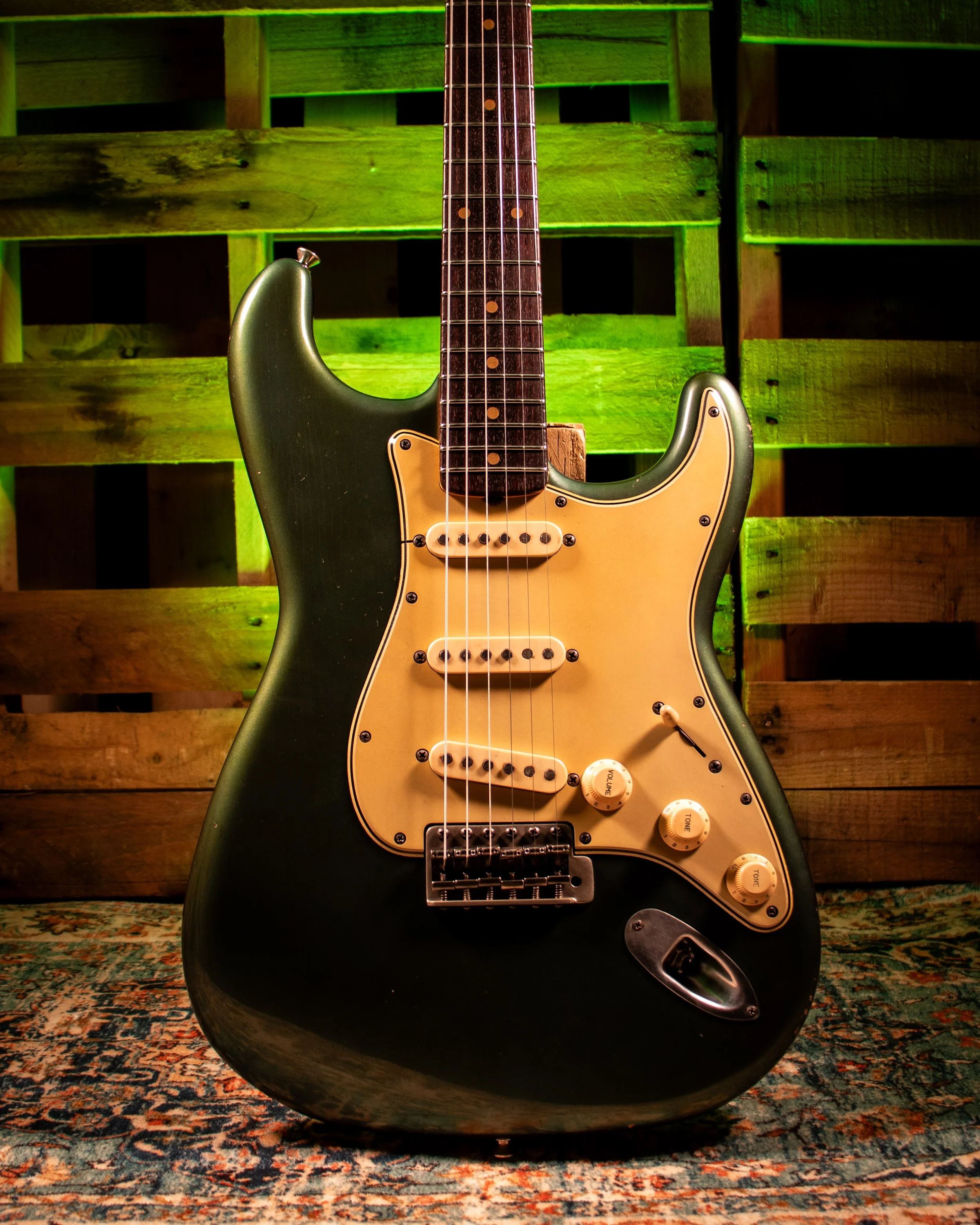 A black electric guitar with a white pickguard, resting on a patterned rug in front of a wooden fence background, illuminated by green lighting.