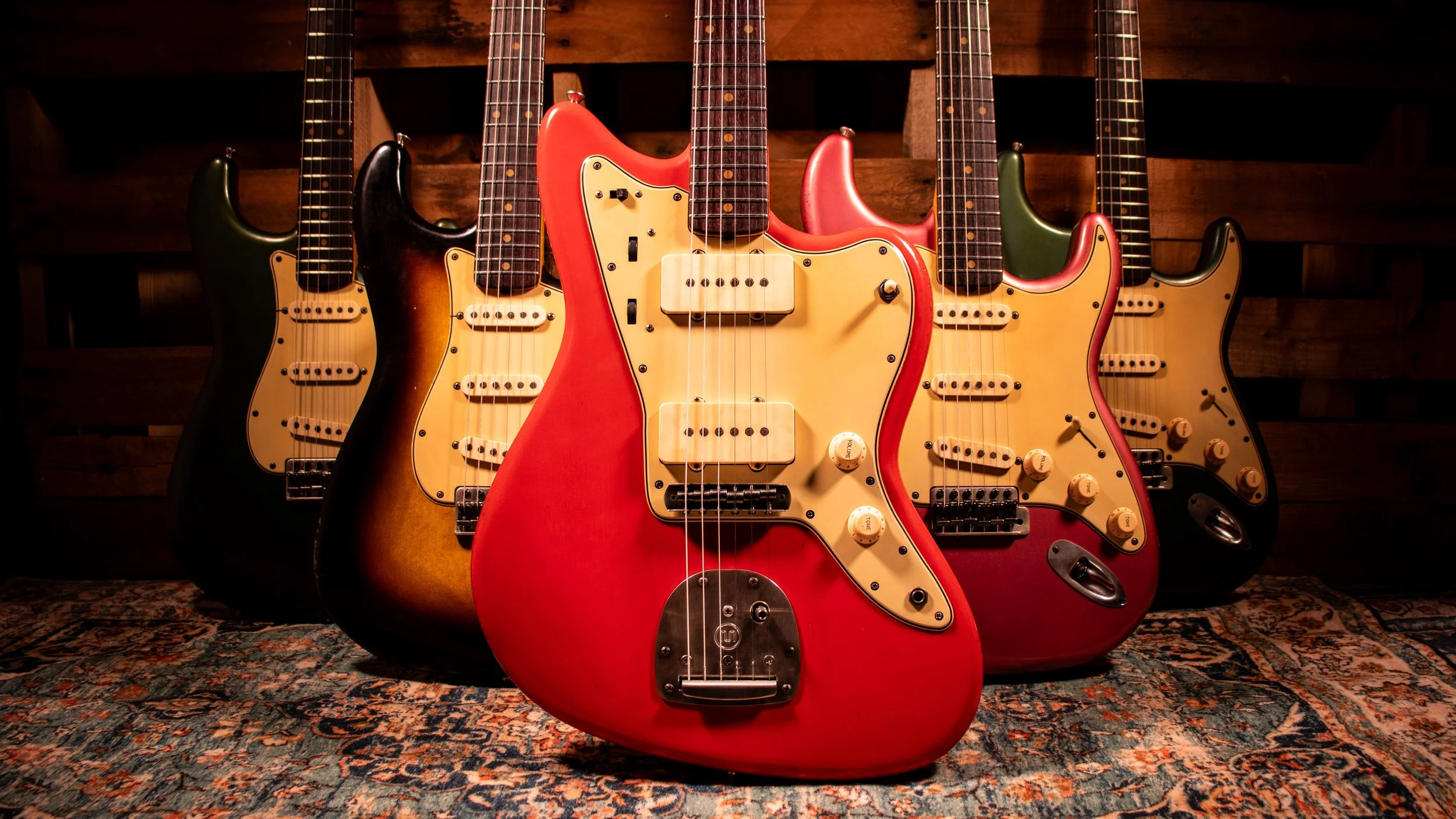 A collection of five electric guitars standing upright on a patterned rug against a wooden wall. The guitars vary in color, with one in red, one in black, one in green, and two in gradient finishes, featuring multiple pickups and control knobs.