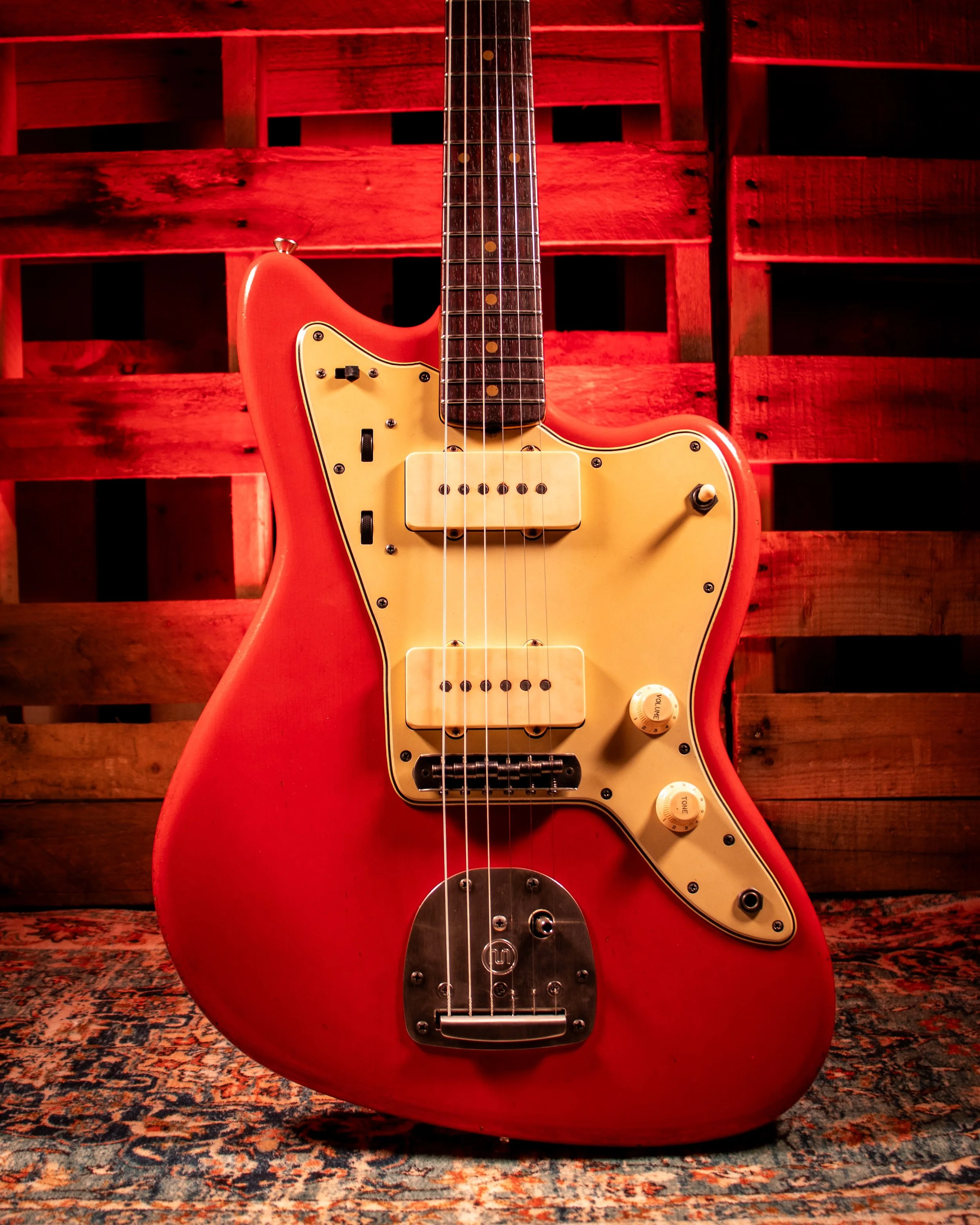 A red electric guitar with a cream pickguard, positioned in front of a wooden backdrop with a patterned rug underneath.
