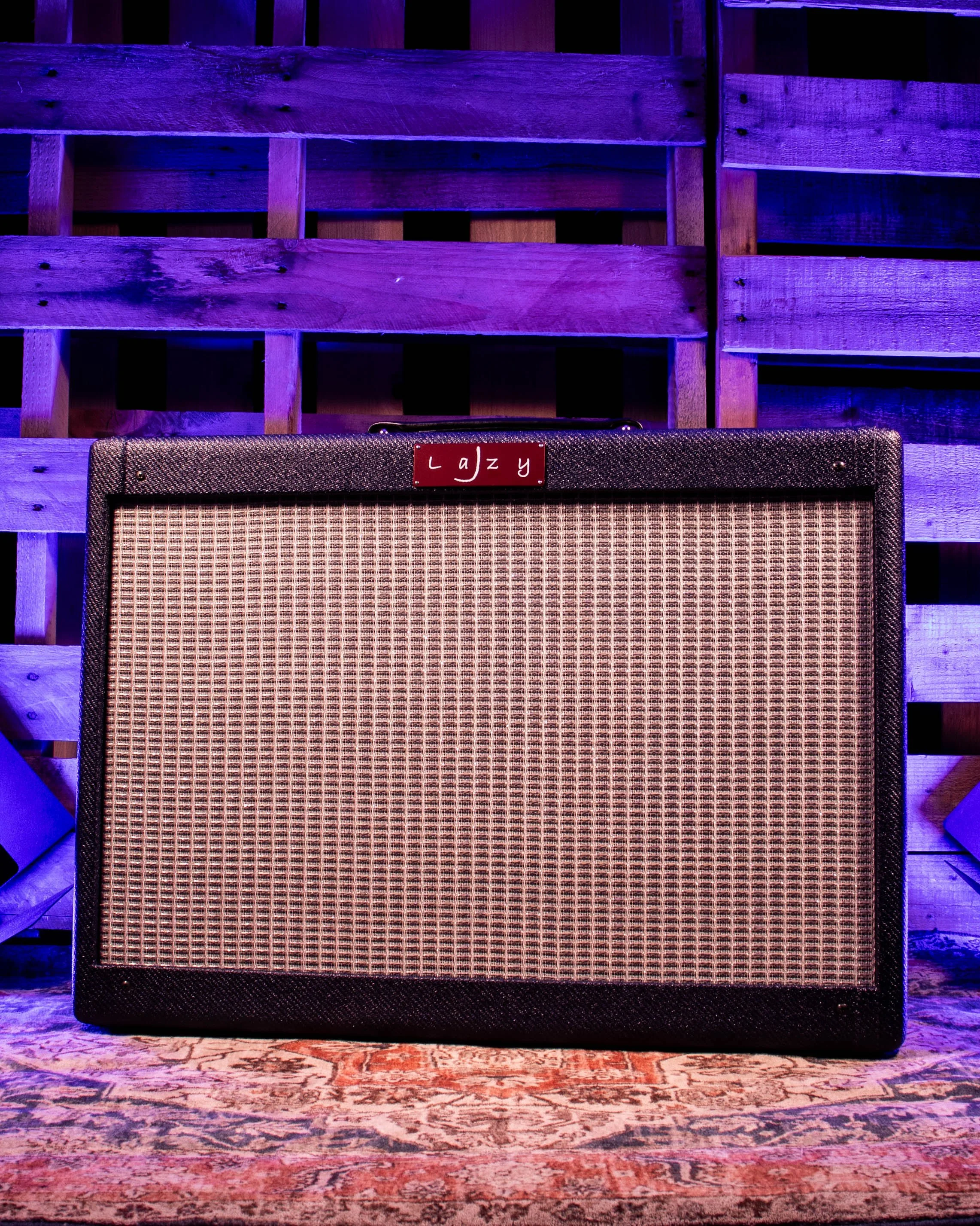 Guitar amplifier with a red nameplate that reads 'Lazy' on a decorative rug with wooden pallets illuminated in purple lighting in the background.