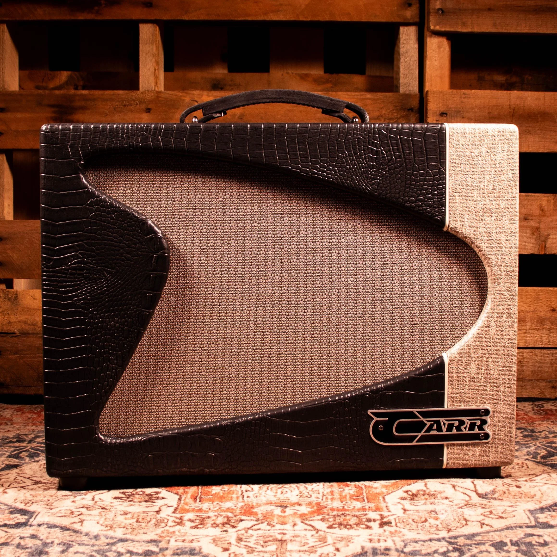 A black and brown crocodile-patterned speaker with a handle, placed on a patterned rug in front of a wooden background.