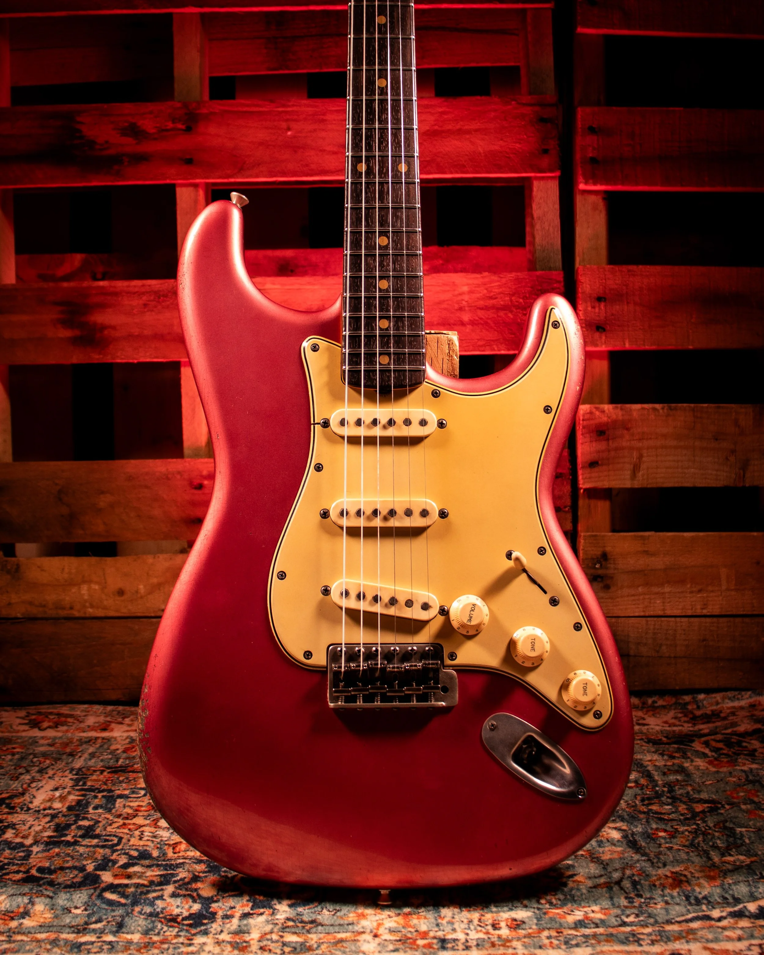 Red electric guitar with a yellow pickguard and control knobs, resting on a patterned rug with a wooden pallet background.