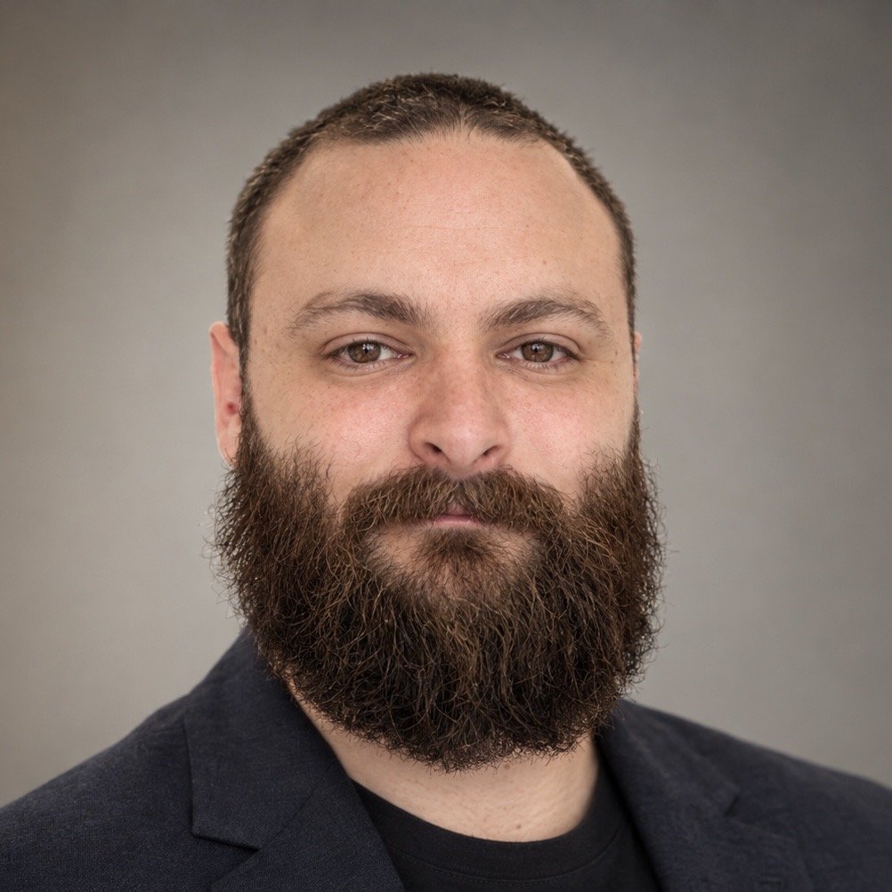 Headshot of a man with a full beard, short brown hair, wearing a dark blazer and black shirt against a neutral background.